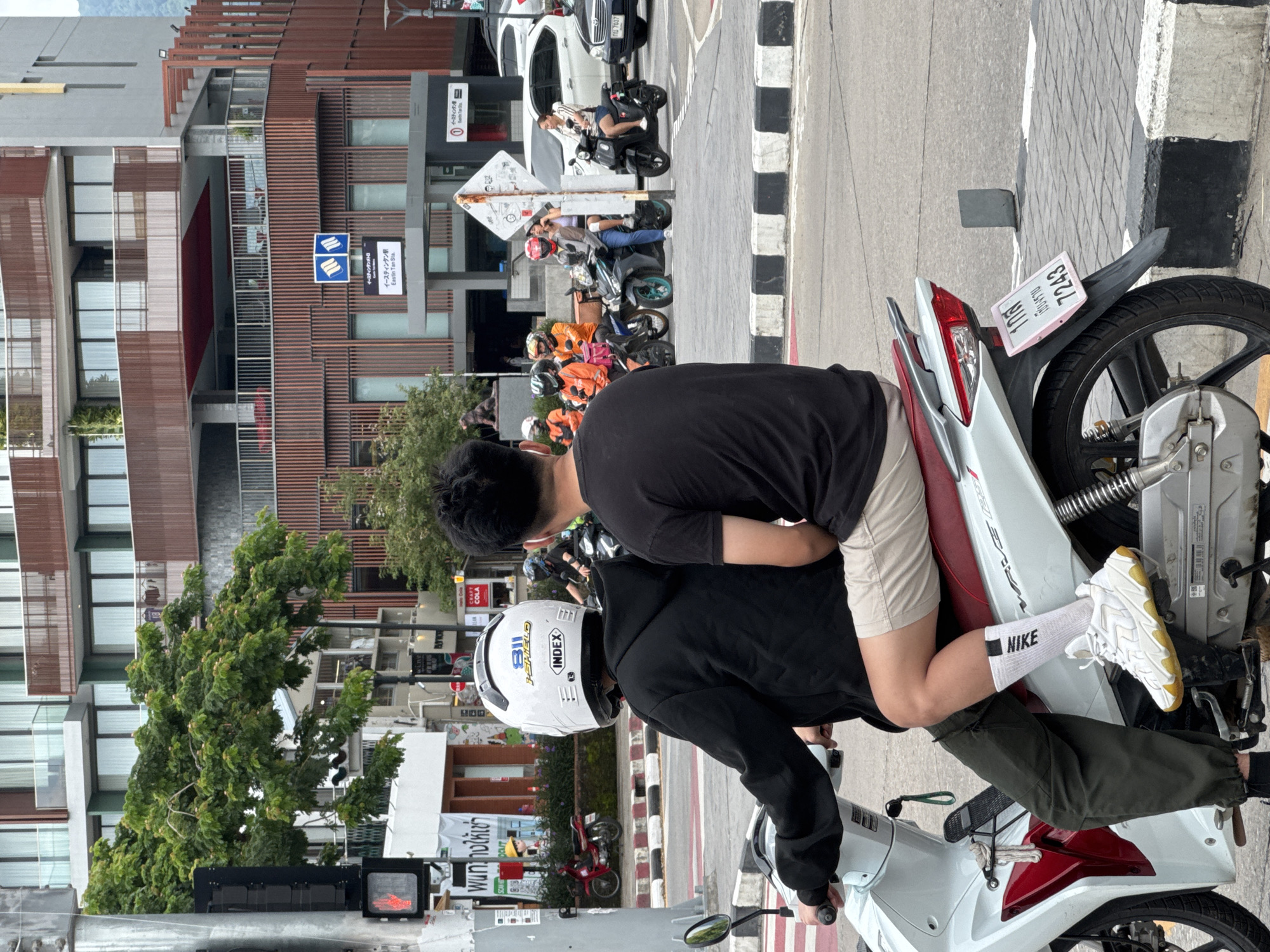 This image captures a street scene in the city of Chiang Mai, Thailand. In the foreground, two individuals are seated on a white and red scooter, with the person in the back wearing a white helmet. The scooter is positioned on a street marked with black and white pedestrian crossings. The background features a modern building with brown and grey facades, balconies, and large windows. Several other motorcycles and a white car are visible on the street, indicating a busy urban environment. A traffic light with a red hand symbol is also present, suggesting a signal for pedestrians to stop. The overall scene depicts a typical urban setting in Chiang Mai, with its characteristic mix of modern architecture and prevalent motorcycle culture.
