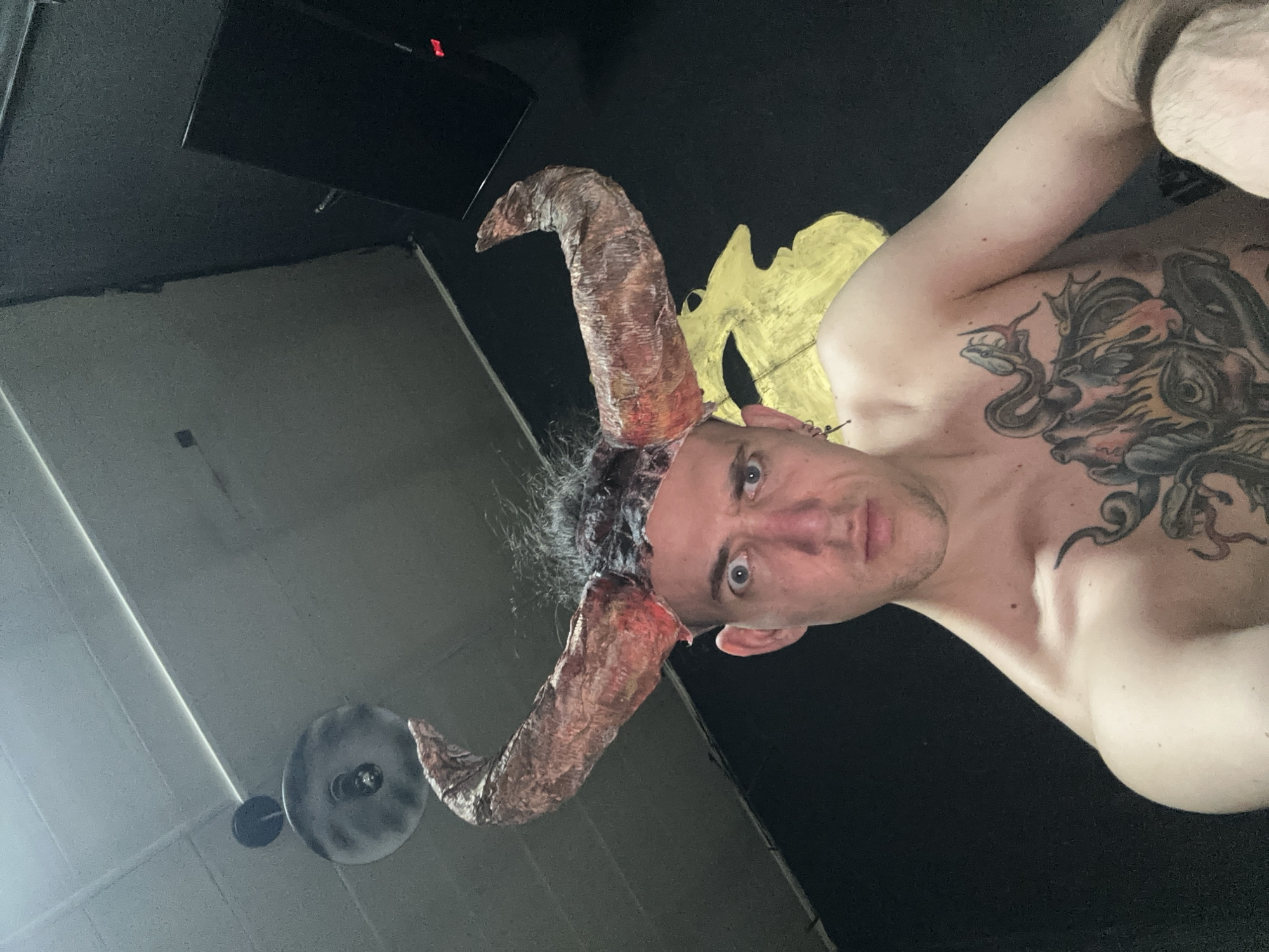 An indoor scene in Saratov, Russia, depicts a shirtless male individual. He wears a costume headpiece featuring two large, curved, textured, reddish-brown horns with dark, scraggly hair. The man looks directly at the camera with wide, light-colored eyes. His chest is heavily tattooed with black and grey designs, including snakes, a winged creature, and an eye. Small silver earrings are visible in his left ear. The background features a light-colored drop ceiling with a dark circular light fixture in the upper left. A dark wall behind the subject displays a yellow, stylized profile painting. In the upper right, a black electronic device with a small red indicator light is present. The environment is dimly lit.