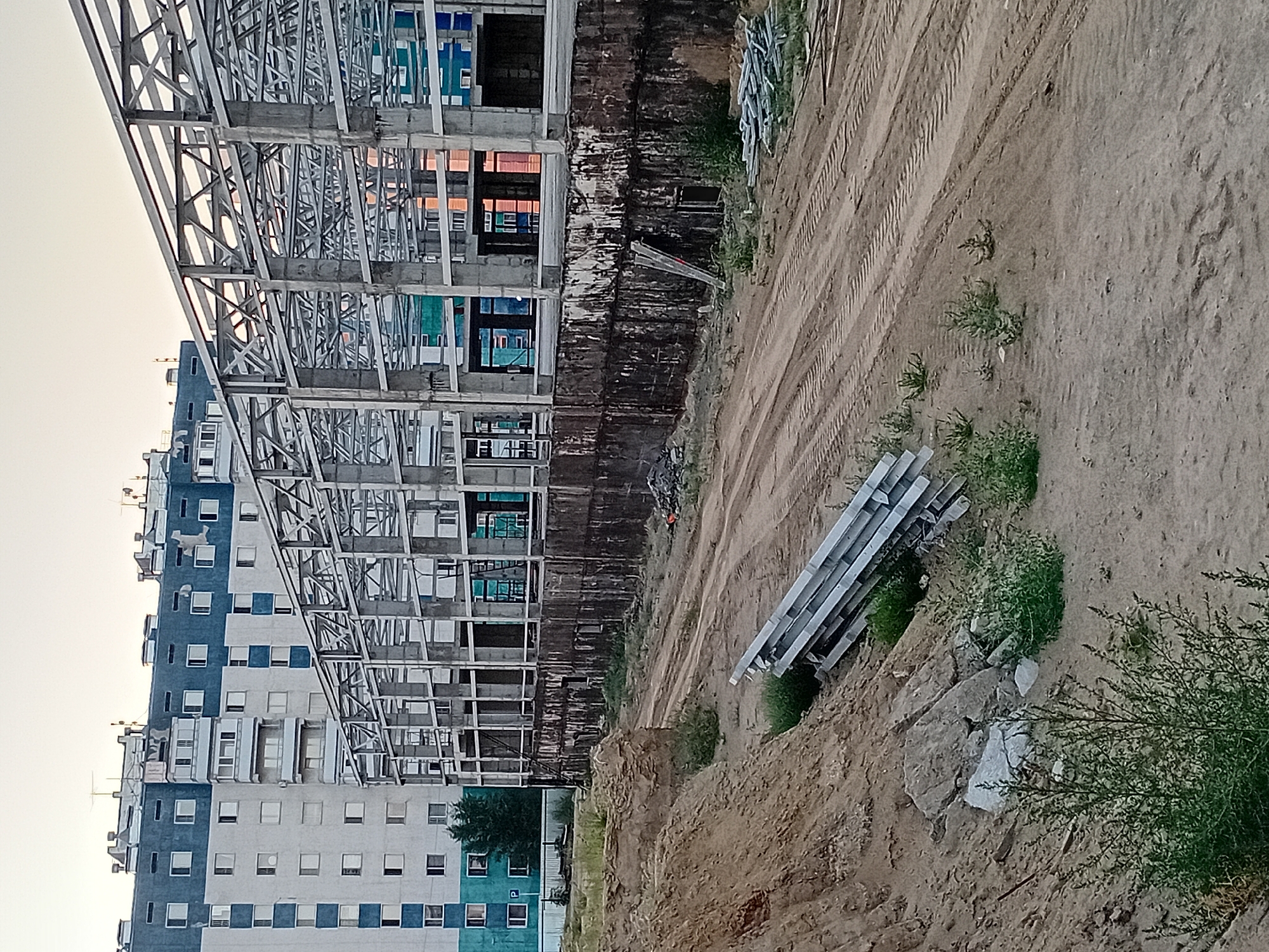 The image depicts a construction site in the city of Semey, Kazakhstan, with an unfinished building structure dominating the scene. In the background, a multi-story residential building with a blue and white facade stands prominently, its windows reflecting the sky. In the foreground, a dirt-covered area shows tire tracks from heavy machinery, indicating ongoing construction work. A pile of metal beams lies on the ground near a mound of dirt and rocks. The overall impression is one of ongoing development and urban expansion.