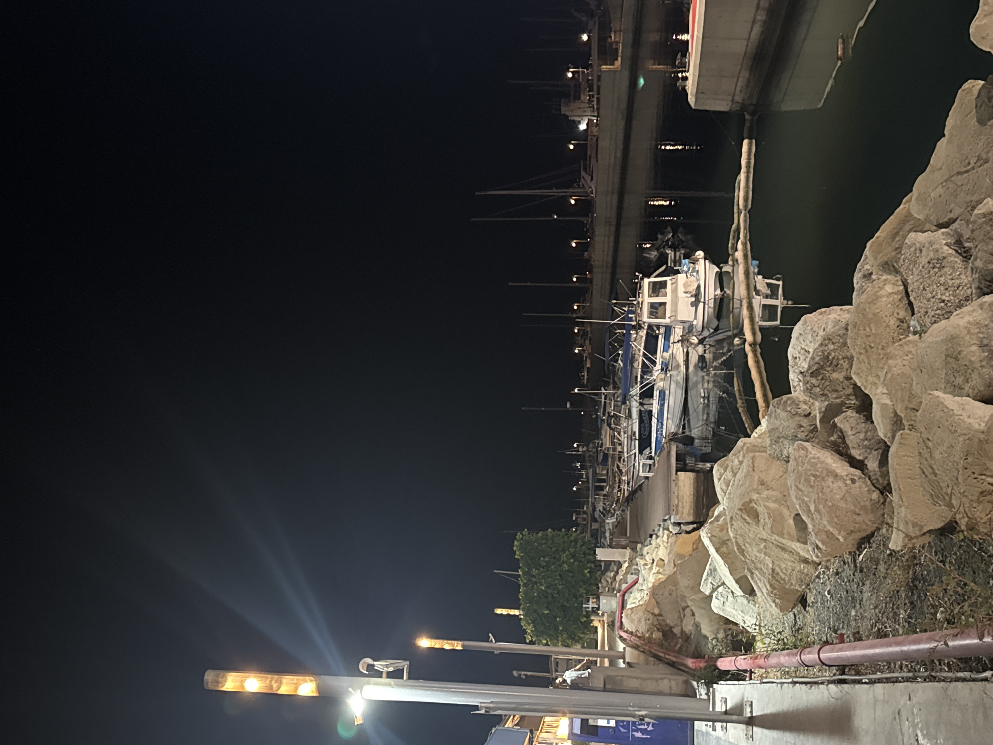 The image depicts a harbor scene at night in Limassol, Cyprus. In the foreground, large rocks line the edge of the water, with a red pipe running along the left side. Further into the scene, several boats are docked in the calm water. The boats vary in size and design, with some appearing to be fishing vessels and others leisure craft. The water reflects the lights from the harbor and the sky, which is dark and clear. Along the waterfront, there are structures that appear to be part of the harbor's infrastructure, including piers and lighting poles. The overall impression is one of a quiet and tranquil night at the harbor, with the artificial lights illuminating the docked boats and the surrounding area.