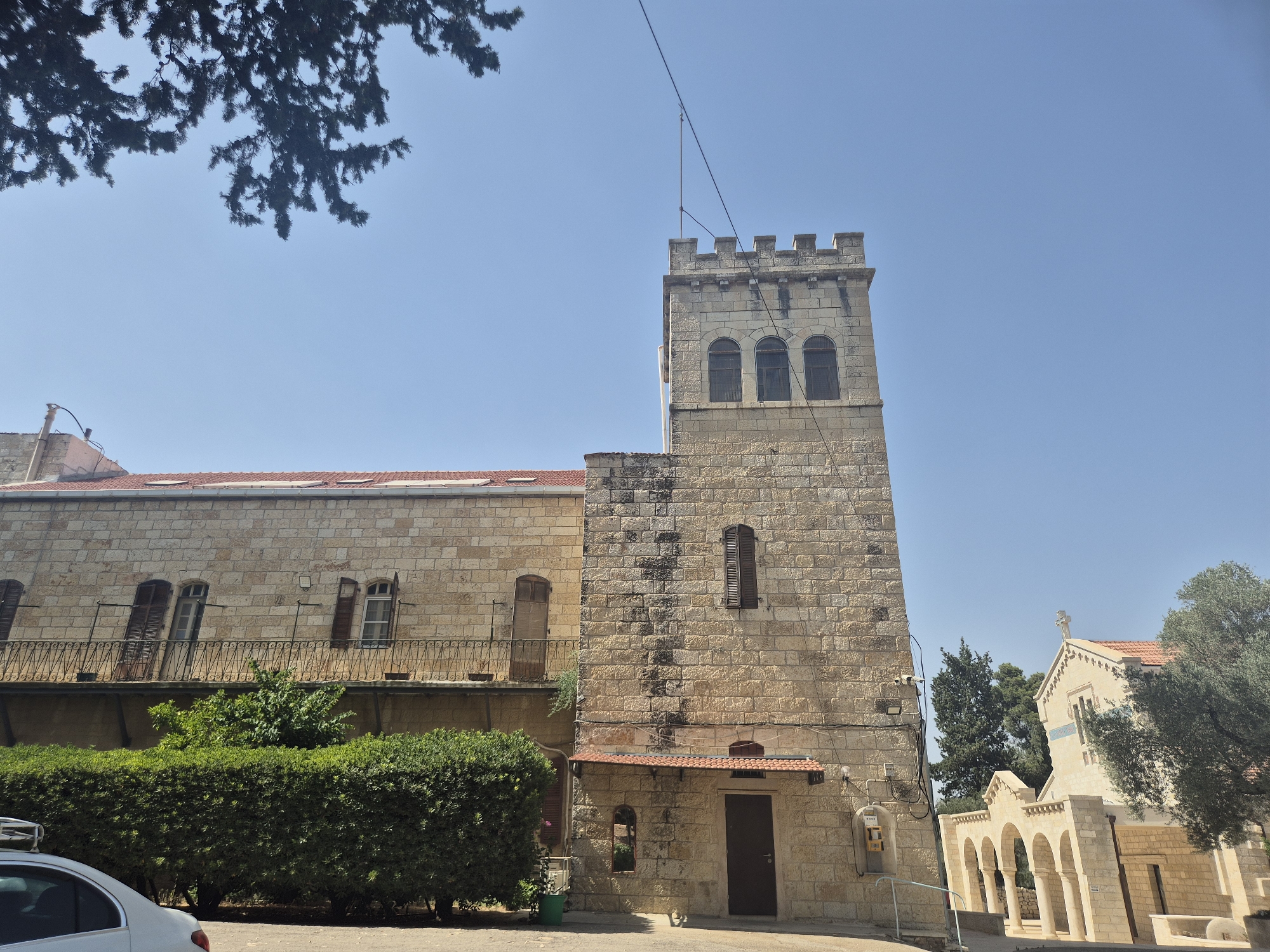 The image depicts a stone building with a tower and a lower wing, situated in Abu Gosh, Israel. The tower, prominently featured on the right, is topped with crenellations and has three arched windows on its upper level. The main building, to the left of the tower, is also constructed of stone and features a red-tiled roof. A balcony with a decorative railing runs along the second story of the main building, above a well-maintained hedge. To the far right, another stone building with arched colonnades and a cross on its roof is visible, suggesting a religious or historical site. A white car is partially visible in the lower left corner. The sky is clear and blue, and tree branches overhang the top left of the frame.