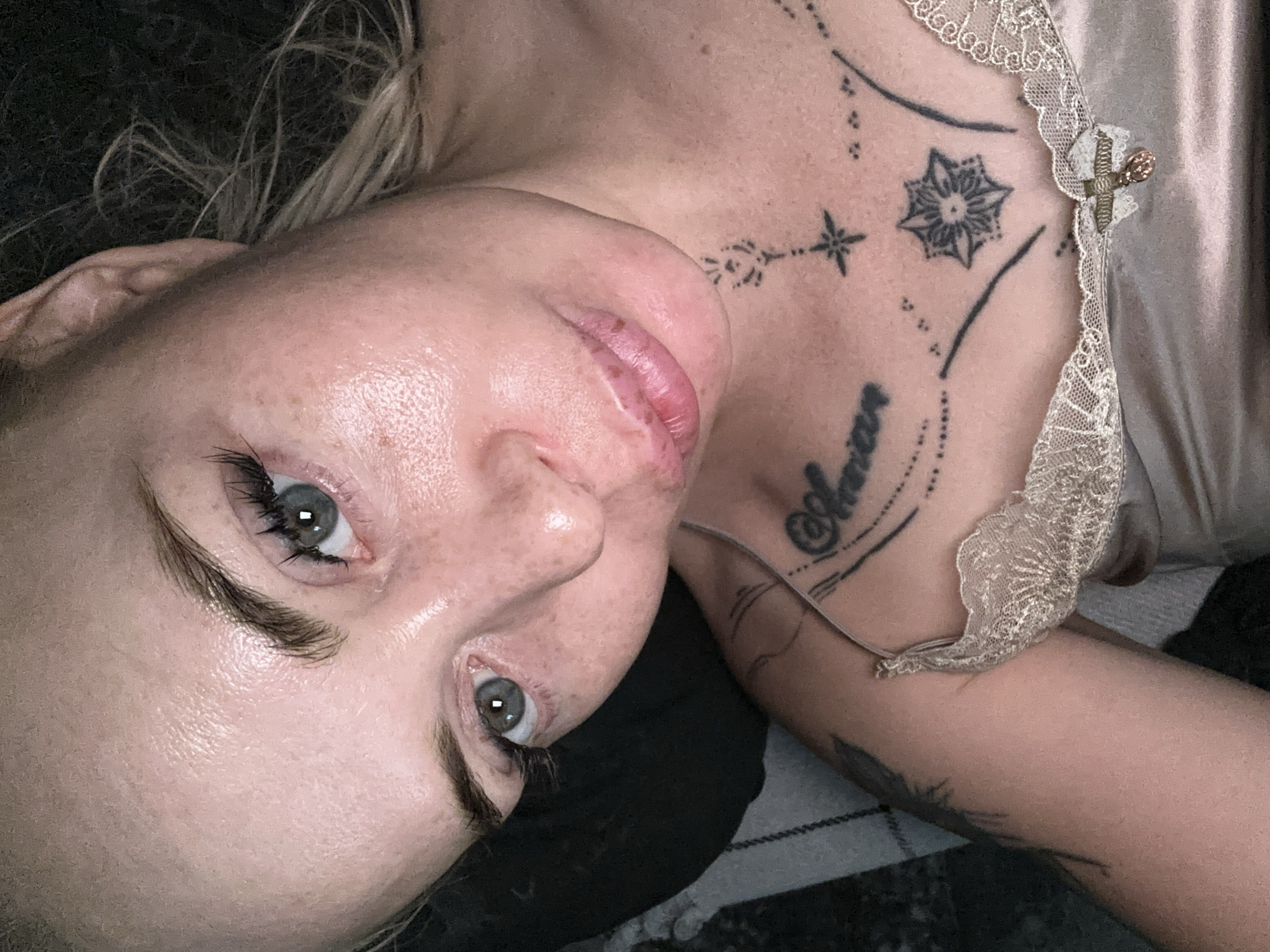 A close-up, overhead shot captures a woman with blonde hair and light-colored eyes. Her skin is lightly freckled, and she is wearing a light-colored satin camisole with lace trim. Several black tattoos are visible on her chest and upper arm, including what appears to be script and geometric designs. The woman's face is tilted slightly upwards, and her lips are slightly parted. The background is dark and appears to be a bedroom setting, with a hint of a patterned fabric on the left side of the frame. The lighting suggests an indoor environment, possibly at night or in a dimly lit room.