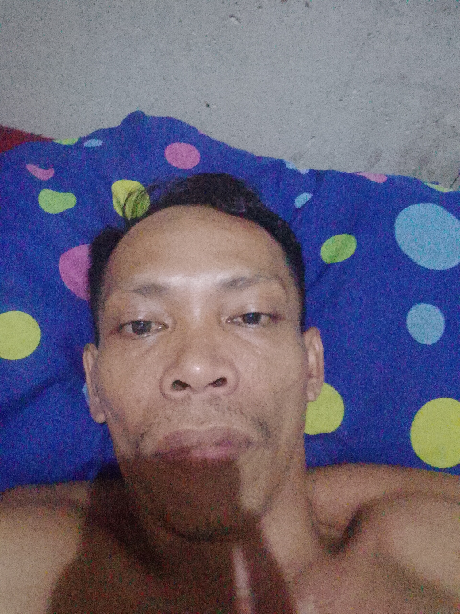 A close-up, eye-level selfie of a man with dark hair and light brown skin, lying on a bed covered with a blue polka-dotted bedsheet (yellow, green, pink and light blue). The location is inferred to be in Bukit Ketok, Indonesia. The man is looking directly at the camera. Part of his bare shoulders are visible in the foreground. The background is slightly out of focus due to the close proximity of the subject. The man has a thin mustache and a neutral expression. The lighting is dim. The ceiling, which appears to be concrete, is partially visible above the bedsheet.