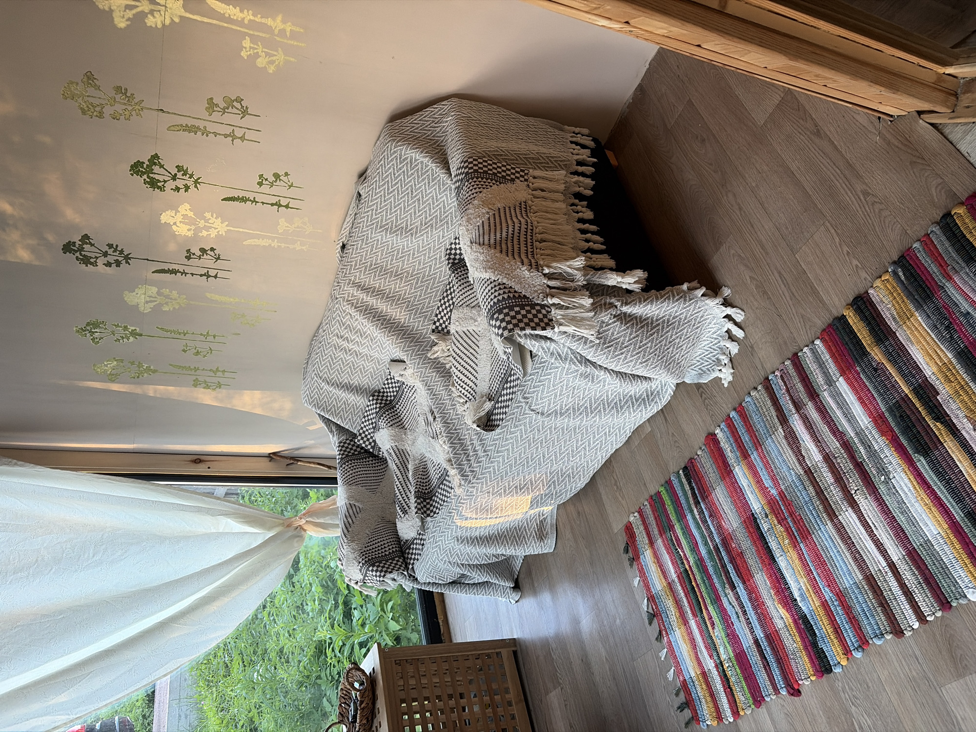 A sunlit interior scene shows a couch draped with patterned throws and blankets, placed in front of a wall adorned with green botanical stencils. A woven rug with colorful stripes lies on the wooden floor in the foreground. To the left, a wooden side table with a basket on top is visible next to a large window with sheer curtains revealing lush green foliage outside. The overall impression is one of a cozy and naturalistic living space, possibly in Snegiryovka, Russia, bathed in the warm glow of natural light. There are no people or animals present.