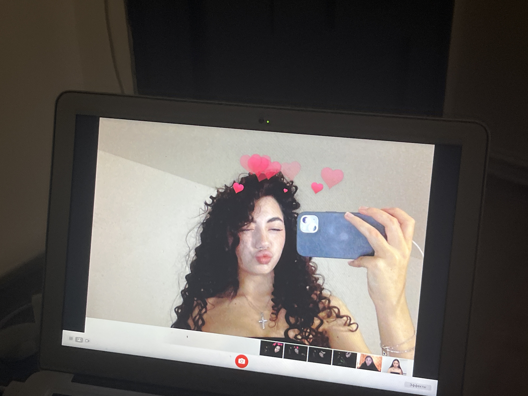The image is a close-up shot of a laptop screen displaying a video call or recording. On the screen, a young woman with long, curly dark hair is taking a selfie with her phone. She is puckering her lips and has a playful expression. Pink heart emojis are overlaid on the image around her head, suggesting a fun or romantic theme. She is wearing a silver cross necklace and a bracelet. The background appears to be a plain wall. A webcam light is visible above her image on the laptop's bezel. The bottom of the screen shows a row of thumbnail images, likely from the video call, and a button labeled "Эффекты," which means "Effects" in Russian, indicating that some form of video filters or effects are being used. The laptop is placed on a surface, and the surrounding environment is dimly lit, with the laptop screen being the primary light source. The image does not contain any information that would suggest a location in the city of Chotěšov, Czechia.