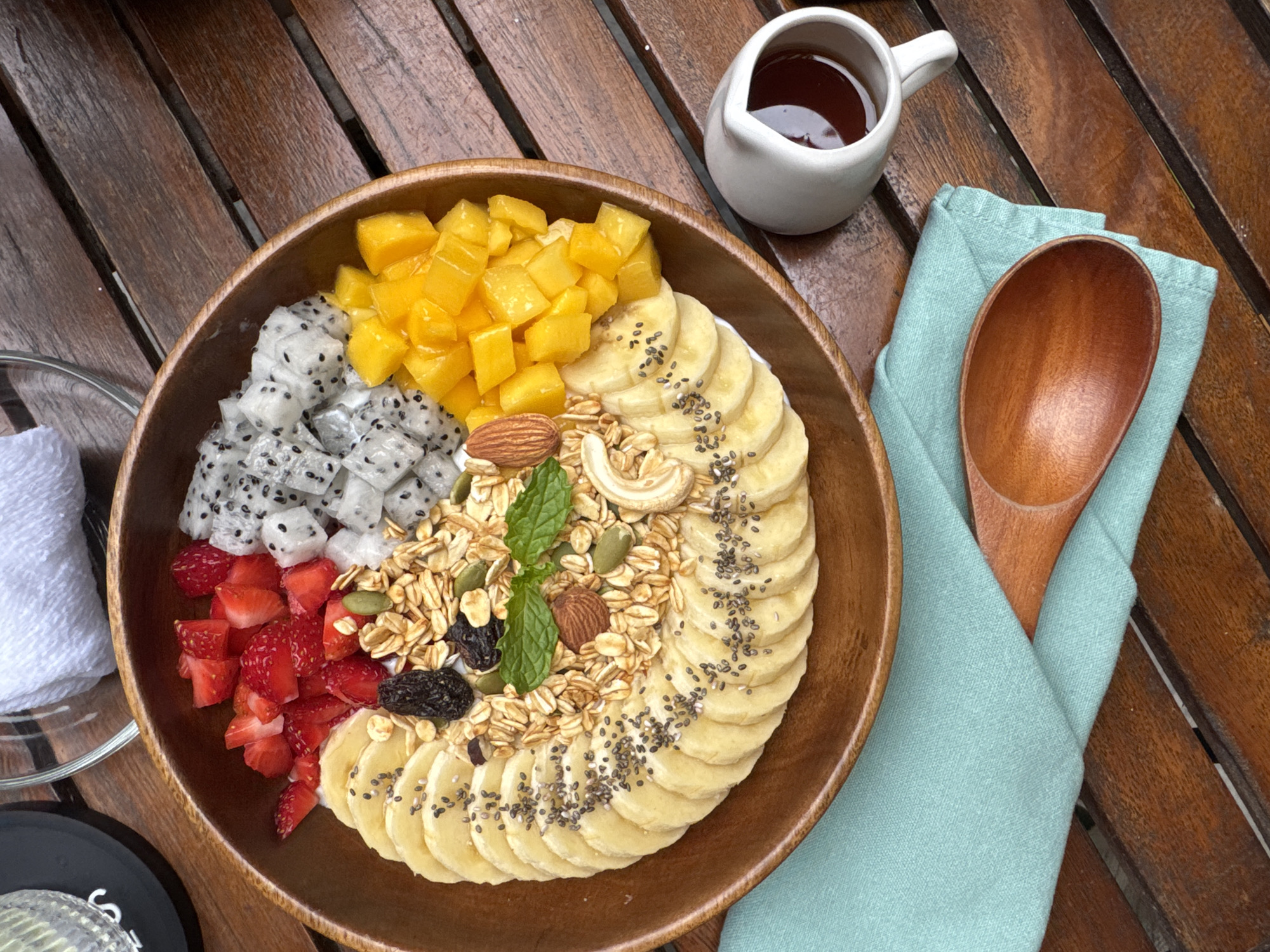 An overhead shot captures a vibrant and healthy smoothie bowl served on a dark wooden slatted table. The main subject is a large wooden bowl filled with a colorful array of fresh fruits, granola, and nuts.
The smoothie bowl features neatly arranged sections of diced mango (bright yellow), cubed dragon fruit with black seeds (white and speckled), and chopped strawberries (red). A large, fan-like arrangement of sliced bananas, sprinkled with black chia seeds, fills another segment of the bowl. The center of the bowl is generously covered with granola, mixed with pumpkin seeds, almonds, cashews, and dark raisins. A few fresh green mint leaves garnish the granola.
Beside the bowl, a small white ceramic pitcher containing a dark liquid, likely syrup or honey, is placed. To the right of the bowl, a light blue linen napkin is folded, with a rustic wooden spoon resting on top of it. In the bottom left corner, part of a small white rolled towel and a glass on a black coaster (with an indistinct partial letter 'S' visible) can be seen.
The scene depicts a ready-to-eat breakfast or brunch, emphasizing fresh, tropical ingredients commonly found in regions like Da Nang, Vietnam. The time of day appears to be morning or early afternoon, given the nature of the meal. The overall impression is one of freshness, health, and a pleasant dining experience in a natural, possibly outdoor, setting.