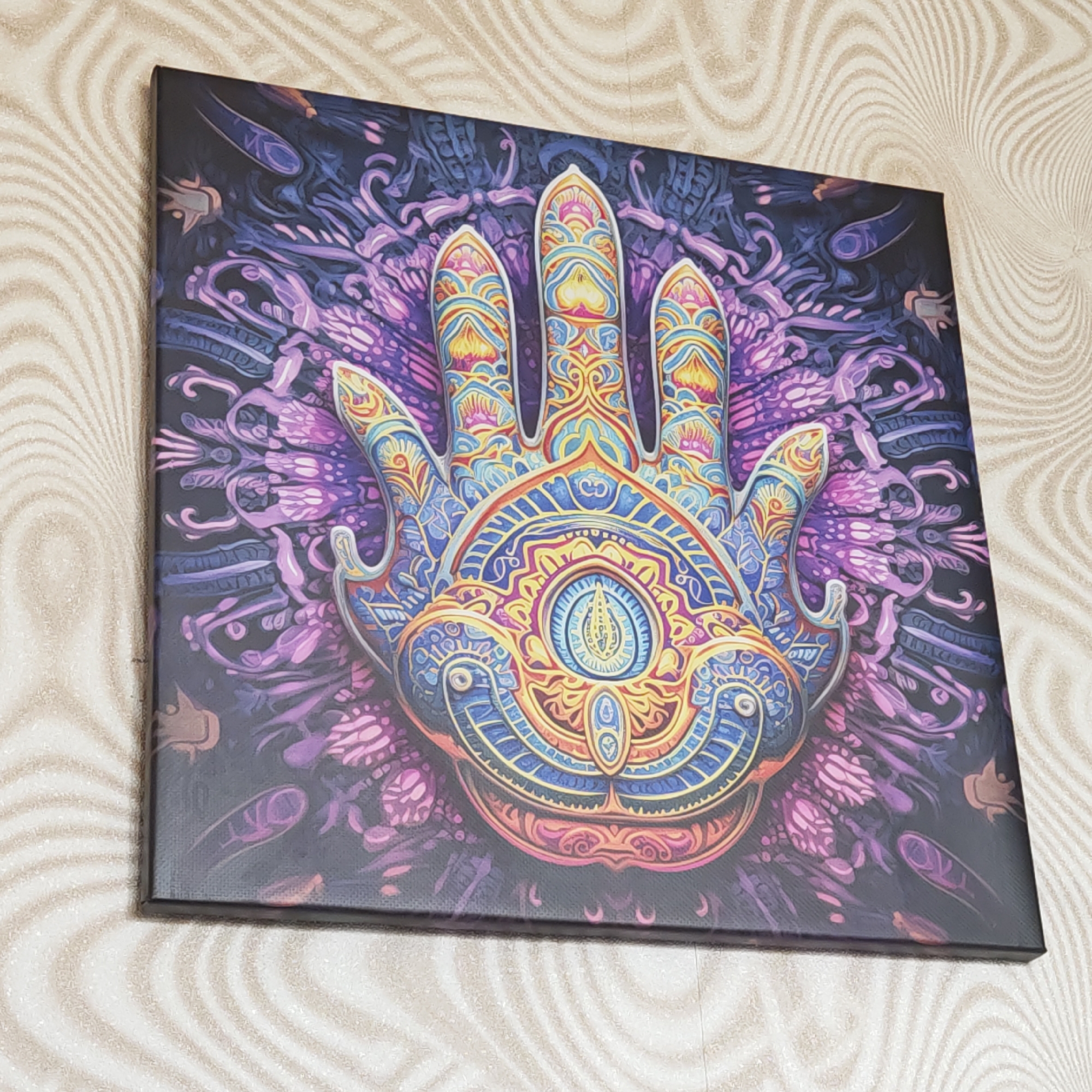 A square canvas print featuring an ornate, colorful depiction of a hand with an eye in its palm is mounted on a wall with a wavy, neutral-toned pattern. The hand is primarily blue, yellow, and orange, with detailed designs on the fingers and palm. The background of the print consists of dark purples and blues with intricate patterns. The location is a room with patterned wallpaper. There is no indication of the city Rostov, Russia.