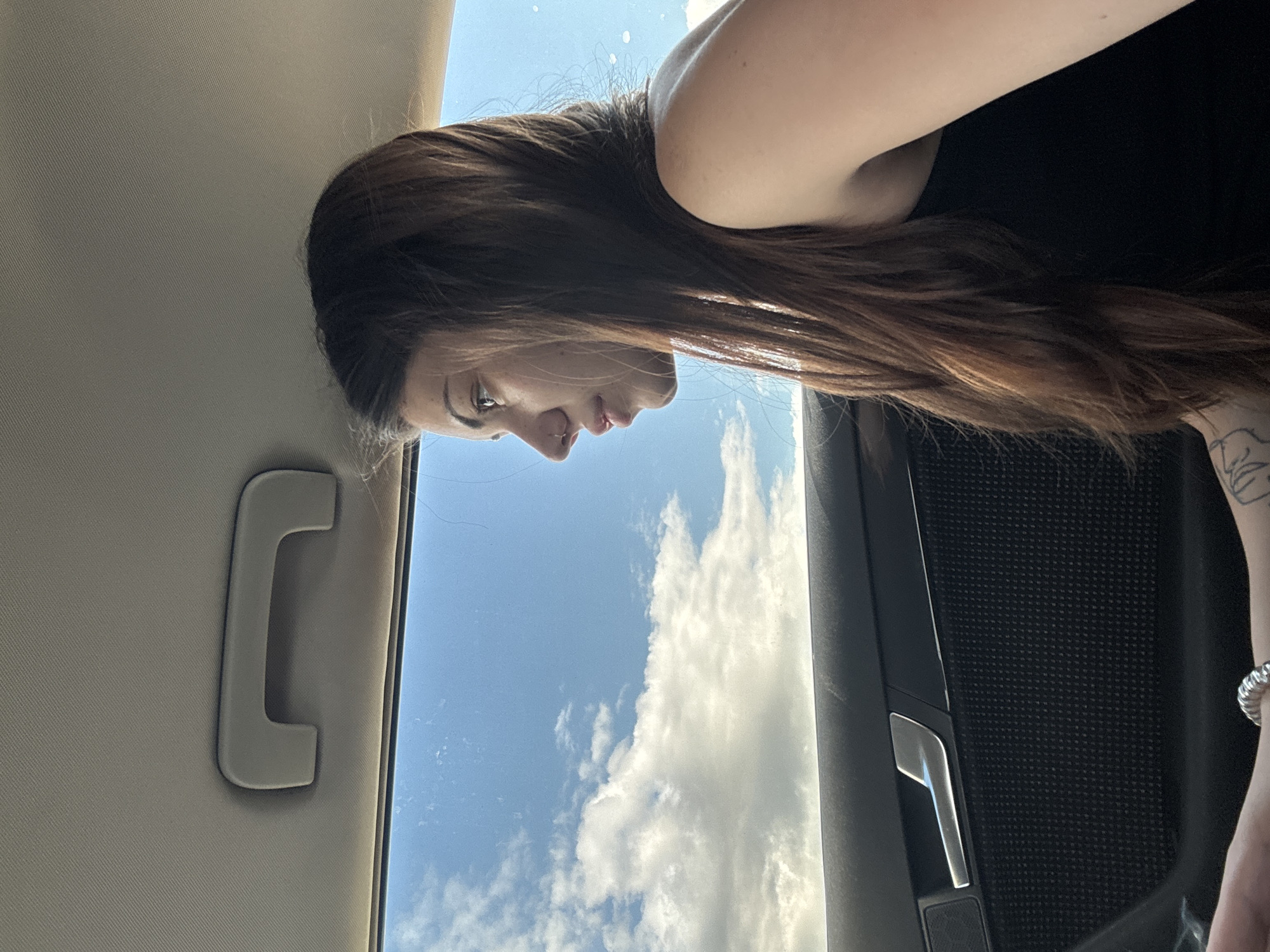 This media file captures a side profile of a woman inside a car, looking out towards a bright sky with clouds. The image is oriented sideways, but if viewed upright, the woman's head is on the right, facing towards the left and slightly downwards. She has long, wavy brown hair. Her face is in profile, revealing a subtle nose piercing. She is wearing a dark top, and a tattoo is visible on her left forearm/wrist area, along with a silver ring on her finger.
The setting is inside a car, with elements of the vehicle framing the outdoor view. On the left side of the image (or top, if upright), a beige car headliner and a matching grab handle are visible. On the right side (or bottom, if upright), part of a dark, textured car door panel with a silver handle is seen. The central portion of the image is dominated by a clear blue sky filled with bright, white, fluffy clouds, indicative of daytime and fair weather. Sunlight is strong, suggesting midday or early afternoon. The woman appears contemplative, observing the sky. The scene is located in Mramor, Bulgaria. No text is visible in the image.