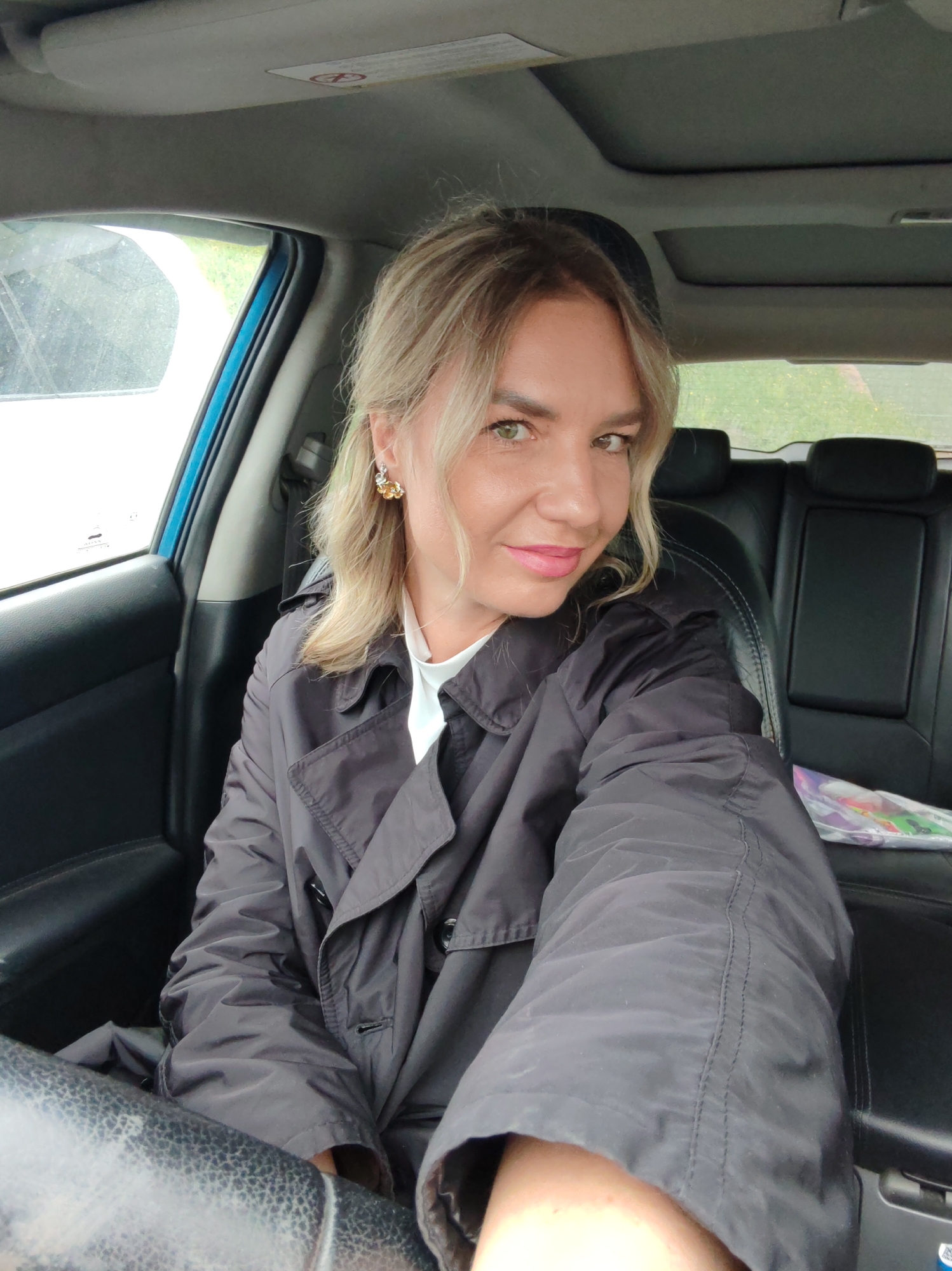 A woman is seated in the driver's seat of a car, looking towards the camera and smiling. She has blonde hair, green eyes, and is wearing a black trench coat over a white top. She is also wearing a gold earring in her left ear. The car interior is visible, with black leather seats and a sunroof. The background shows a glimpse of the outdoors, with greenery and a cloudy sky.