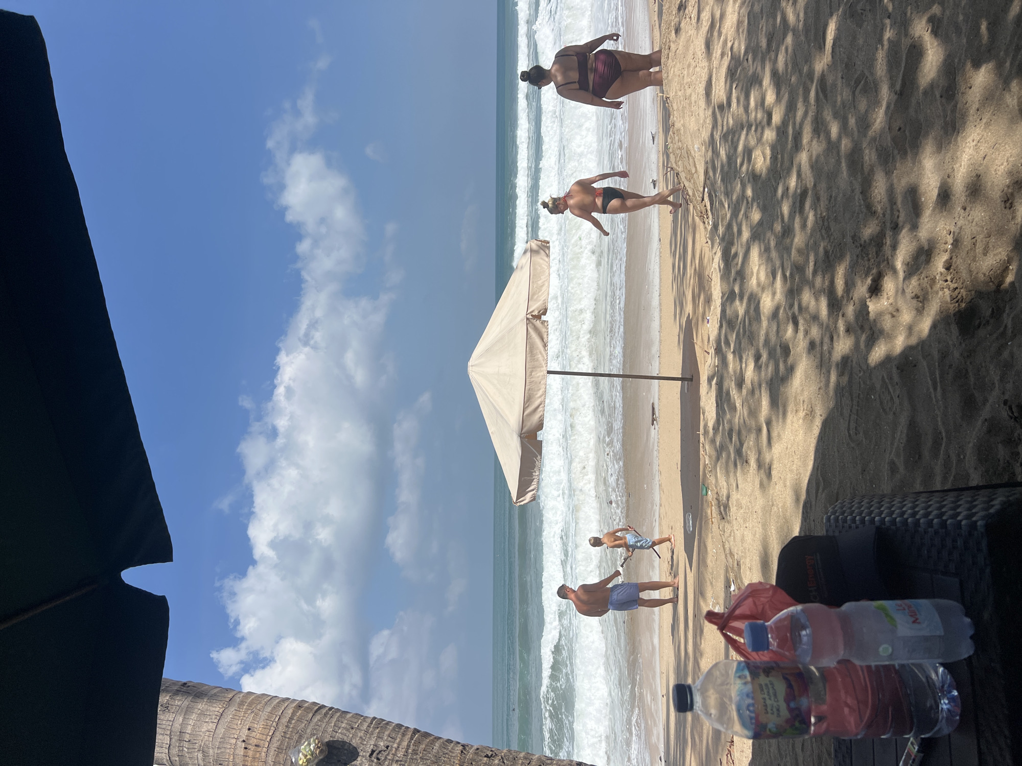 The image depicts a sunny day at Kuta Beach, Indonesia. In the foreground, partially obscured by shadows from palm trees and an umbrella, are two plastic water bottles and a red plastic bag on a dark woven surface. The middle ground shows a sandy beach with the ocean in the background. Waves are breaking on the shore. A man in blue swim trunks stands near the water, facing away from the camera, while a child in similar attire holds a stick. Further down the beach, two women in swimsuits are walking away from the camera towards the ocean, one in a black bikini and the other in a burgundy bikini. The sky is blue with scattered white clouds.