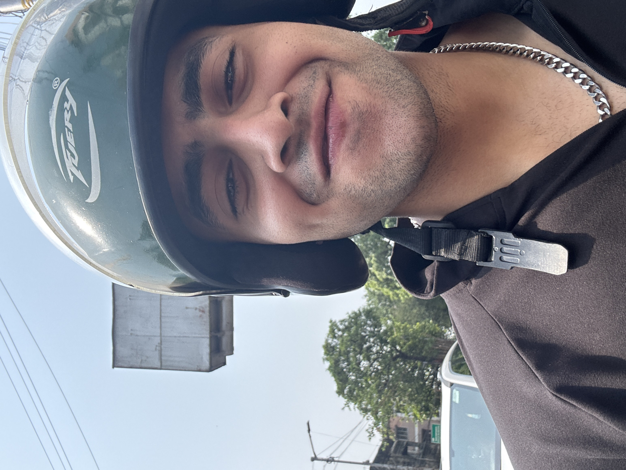 A young man is photographed close-up wearing a light green motorcycle helmet with the word "TIERY" in white letters. He is also wearing a black jacket and a silver chain necklace. The helmet strap is fastened. The man has short dark hair and a light beard. He is smiling slightly. The background is a cityscape, including a power transformer, trees, and buildings. The location is Kolkata, India.