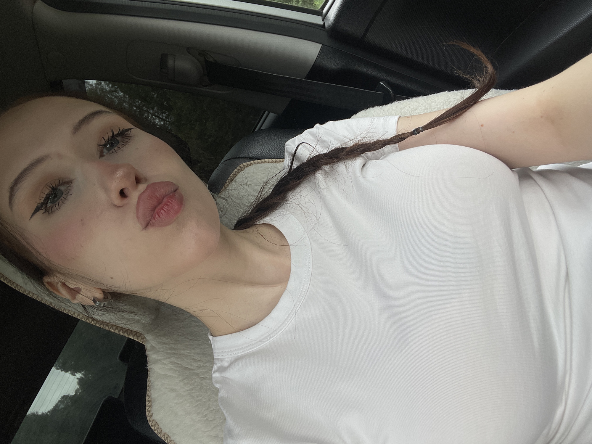 A young woman is taking a selfie in the passenger seat of a car. She has on a white t-shirt and her hair is in a braid. The woman is looking at the camera and making a kissing face. Her makeup is done with eyeliner, eyeshadow, and blush. The car appears to be in a rural area, possibly in Gavrilovka, Russia, as indicated by the trees and greenery visible through the car windows. The lighting suggests it is daytime, possibly overcast, as the light is diffused.