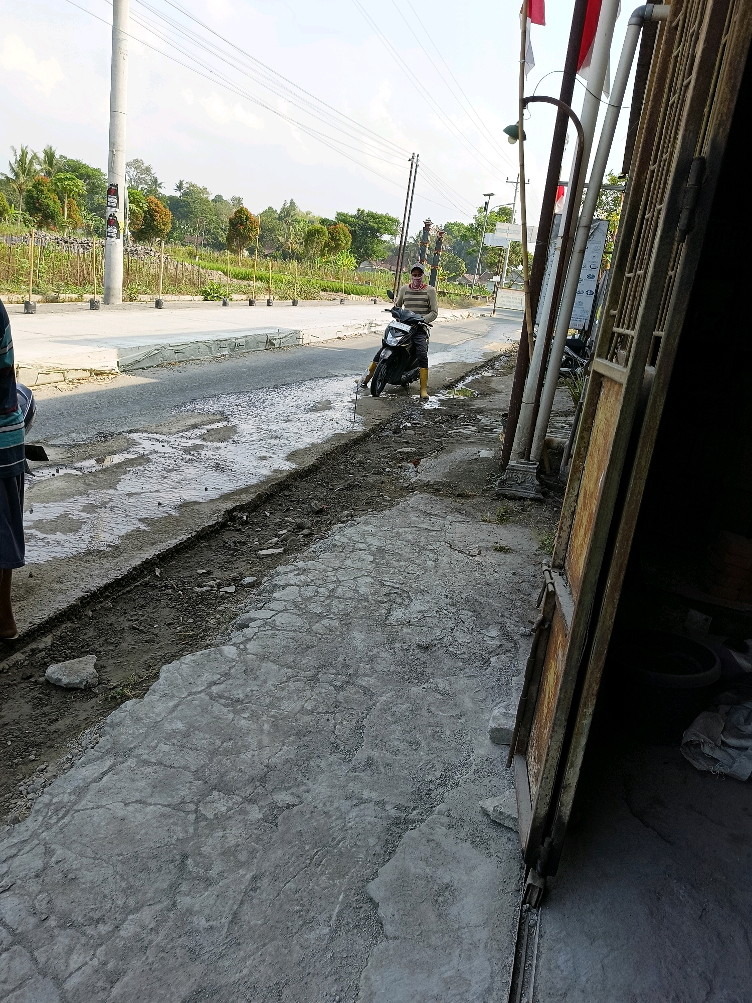 A man on a scooter is parked on the side of a paved road in Sukorini, Indonesia. The road appears to be undergoing some form of repair, with a section of the asphalt removed and the underlying dirt exposed. The man is wearing a striped long-sleeved shirt, dark pants, and yellow boots. He is looking down at the scooter. To the right of the road is a fenced area with what looks like a building and a metal gate. The environment is rural, with greenery and trees visible in the background. The sky is clear with some clouds.