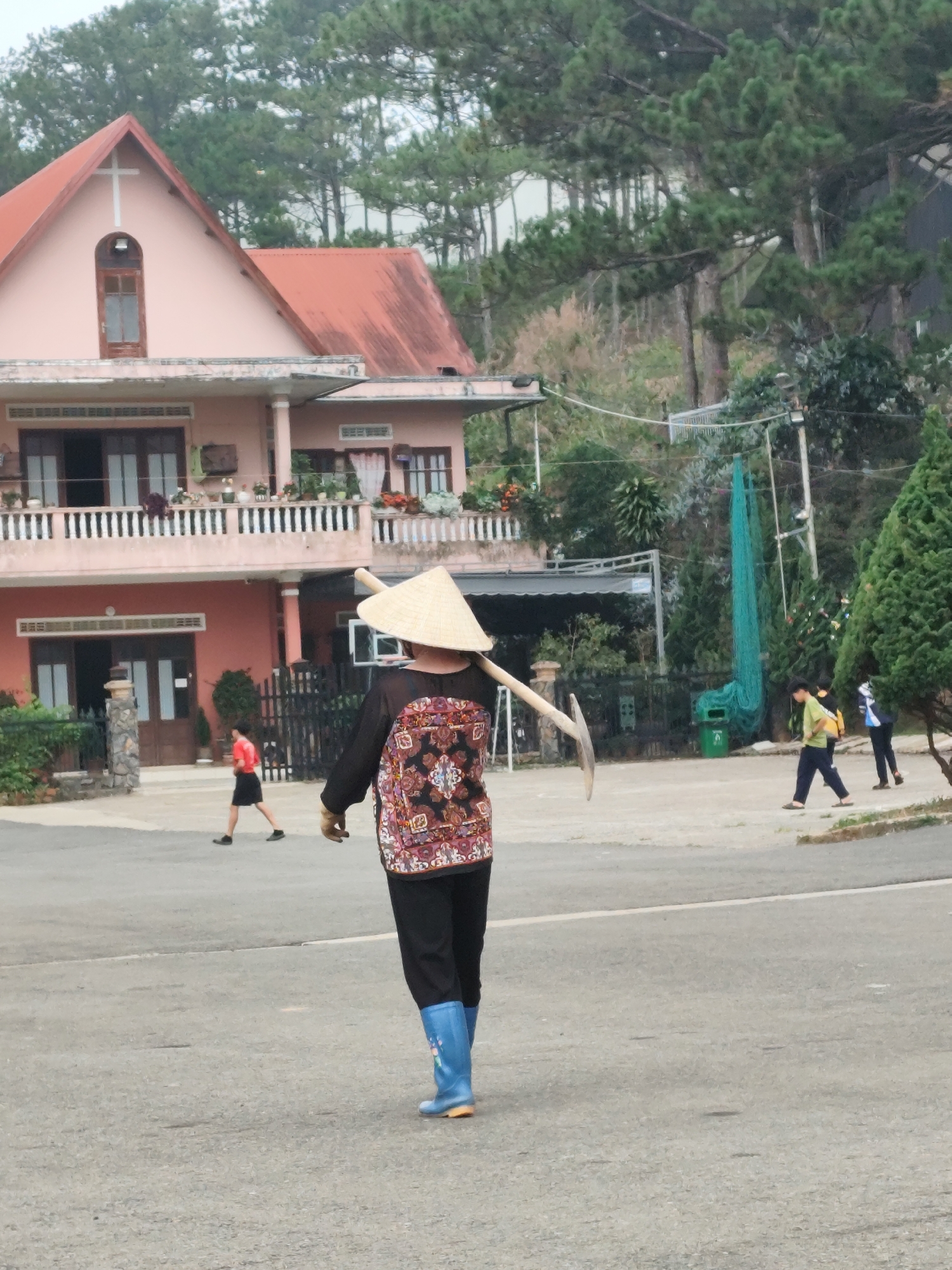 The image portrays a daytime scene in what appears to be a rural or semi-rural area of Lang Biang - Đà Lạt, Vietnam. The central figure, viewed from behind, is a person wearing a traditional Vietnamese conical hat (nón lá), a dark long-sleeved shirt with a vibrant, multi-colored patterned back, black pants, and distinct bright blue rubber boots adorned with a light-colored design. This individual is carrying a long-handled agricultural tool, possibly a hoe or a pickaxe, over their left shoulder, suggesting involvement in farming or gardening work.
In the mid-ground, a prominent two-story house with light peach-colored walls and a reddish-brown tiled roof stands out. The house features a distinctive gable with a white cross at its peak and a narrow arched window below it, along with a balcony on the second floor lined with a white balustrade.
The background is characterized by a lush hillside densely covered with tall pine trees, indicative of a mountainous or hilly landscape. Towards the right side of the image, a long, vertically hanging green mesh net is visible on the hillside, along with a small green trash bin and a black metal fence.
Other people are also present: a person in a red shirt and black shorts walks away from the camera on the left side of the road, and two other individuals are seen walking further down the road on the right.
The overall atmosphere is calm and serene, with soft, diffused daylight suggesting either an overcast sky or an early morning/late afternoon setting. No discernible text is visible in the image. The attire and tools of the main subject, combined with the landscape and architecture, firmly place the scene in rural Vietnam.