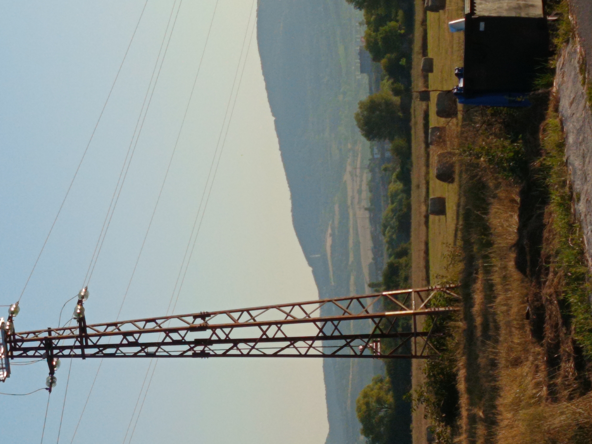 An outdoor, eye-level shot captures a rural landscape bathed in the warm light of what appears to be late afternoon or early morning. Dominating the left side of the frame is a tall, red metal electricity pylon, its lattice structure stark against a pale blue sky. Several wires extend horizontally from the pylon. In the middle ground, a field of dry, golden grass is dotted with several round hay bales, suggesting agricultural activity. To the right, a dark, rectangular dumpster-like container is visible, with a blue tarp draped over it and a white object, possibly a bucket, resting on top. The background features rolling hills covered in vegetation, with a hazy, distant mountainous silhouette forming the horizon. A few trees are scattered throughout the scene, adding patches of green to the predominantly earthy tones. The overall impression is one of a tranquil, rural setting, likely on the outskirts of a town or village. While the exact location is not explicitly stated within the image, the description mentions the city of Kralev Dol, Bulgaria.