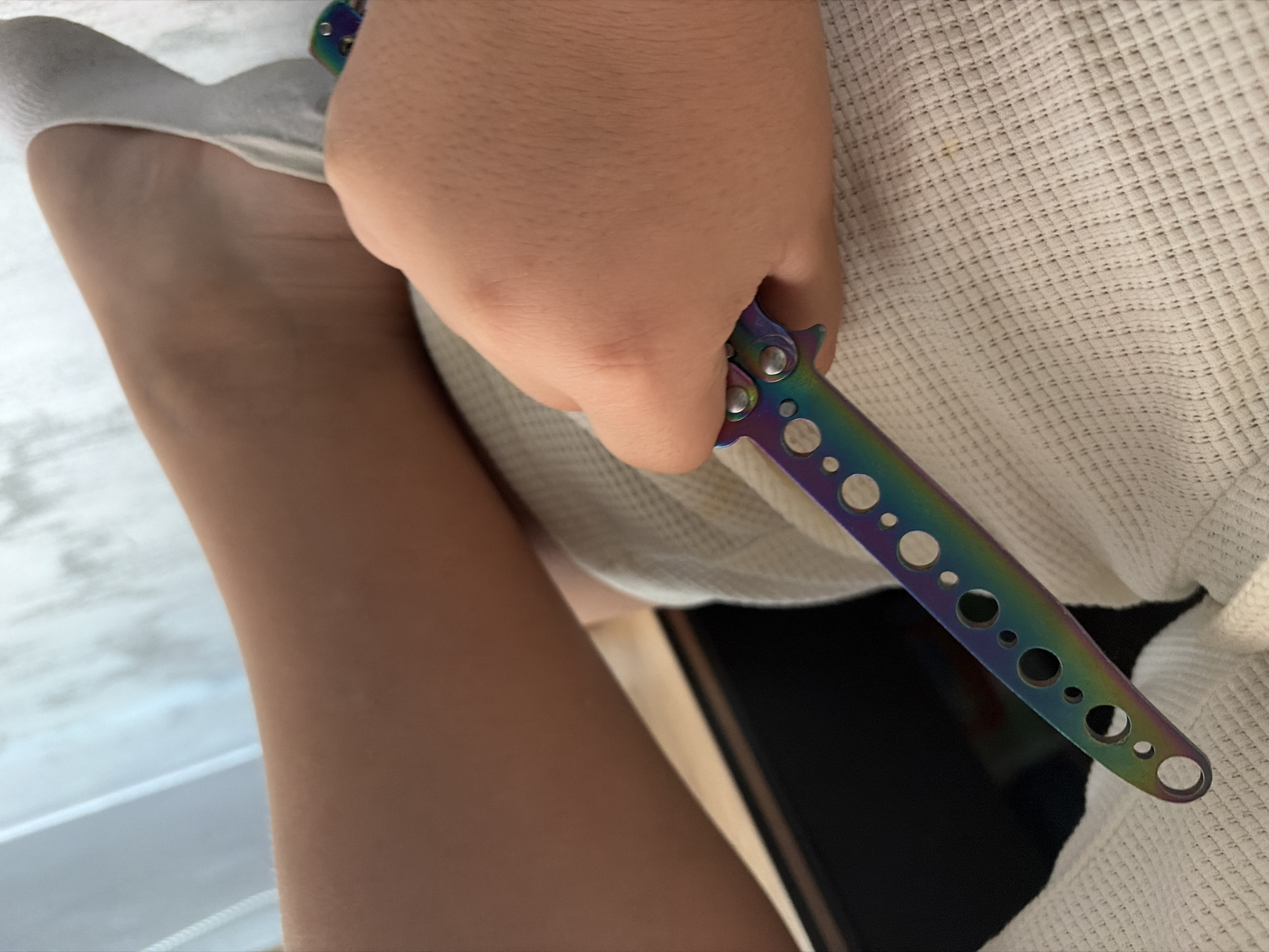 A hand holds a colorful butterfly knife, with a leg and foot visible next to a white textured surface. The setting appears to be indoors, potentially in a private residence. There are no other discernible objects or people. The location in the city of Shumen, Bulgaria cannot be determined from the image.