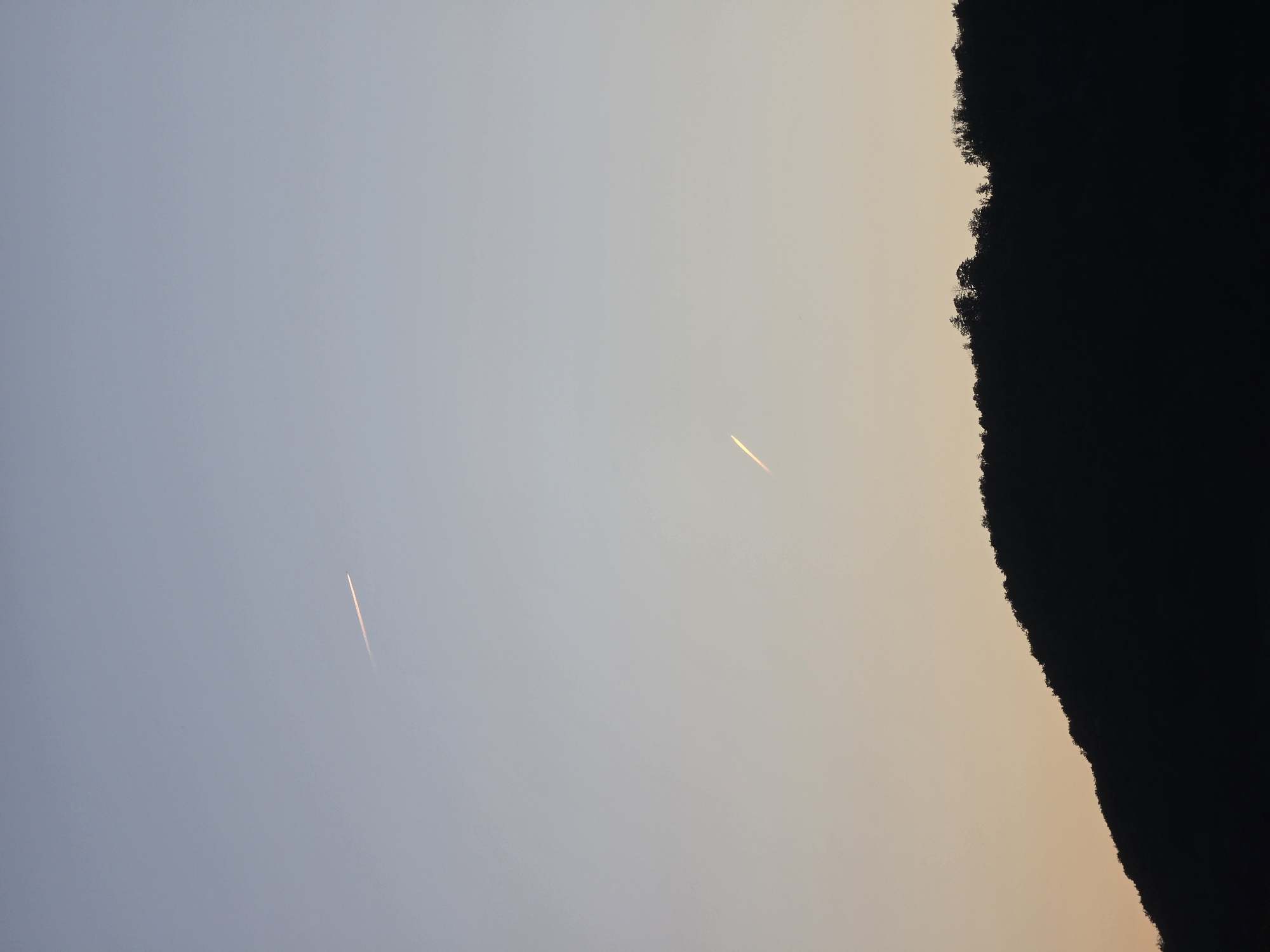 The image depicts a tranquil sunset scene over the city of Godelheim, Germany. In the foreground, the dark silhouette of a treeline outlines a gently sloping hill against the horizon. Above the treeline, the sky transitions from a warm, peachy hue near the horizon to a pale, serene blue at the top of the frame. Two faint contrails, left by airplanes, streak across the sky, adding a subtle dynamic element to the otherwise still atmosphere. One contrail is positioned in the upper portion of the sky, while the second is located lower, closer to the horizon. The overall impression is one of peace and natural beauty as the day gives way to evening in Godelheim.