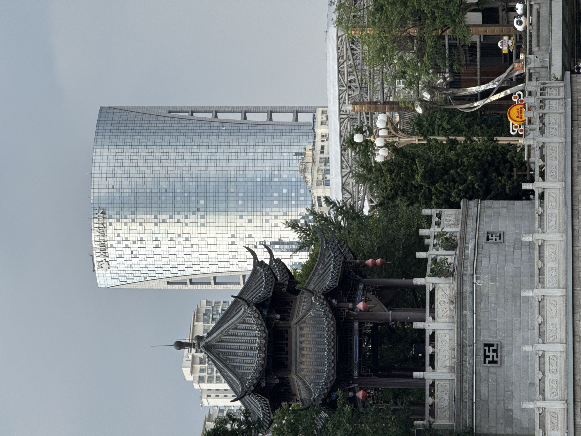 The image presents a blend of traditional and modern architecture in Wuhou District, China, under an overcast sky. In the foreground, a dark, two-tiered traditional Chinese pavilion, featuring a tiled roof with upturned eaves and decorative elements, is situated on a multi-level stone base. Red lanterns hang from the pavilion's upper tier. A person is faintly discernible inside the pavilion. The stone base includes a carved balustrade and fenestration with traditional geometric patterns. To the right, an ornate street light with multiple white globe fixtures rises from a stone railing. Below it, on what appears to be a bridge or waterfront, an abstract metallic sculpture stands alongside a yellow and red sign displaying the characters for "社会主义核心价值观" (Socialist Core Values), and two small panda statues. In the background, a prominent, tall, modern skyscraper with a curved glass facade and vertical architectural cutouts is visible, featuring the "MINYOUN" logo at its top. Several lighter-colored residential buildings and a large, grey, open-truss metal structure are also present in the mid-background. Green trees are dispersed across the scene, providing a natural contrast to the urban structures.