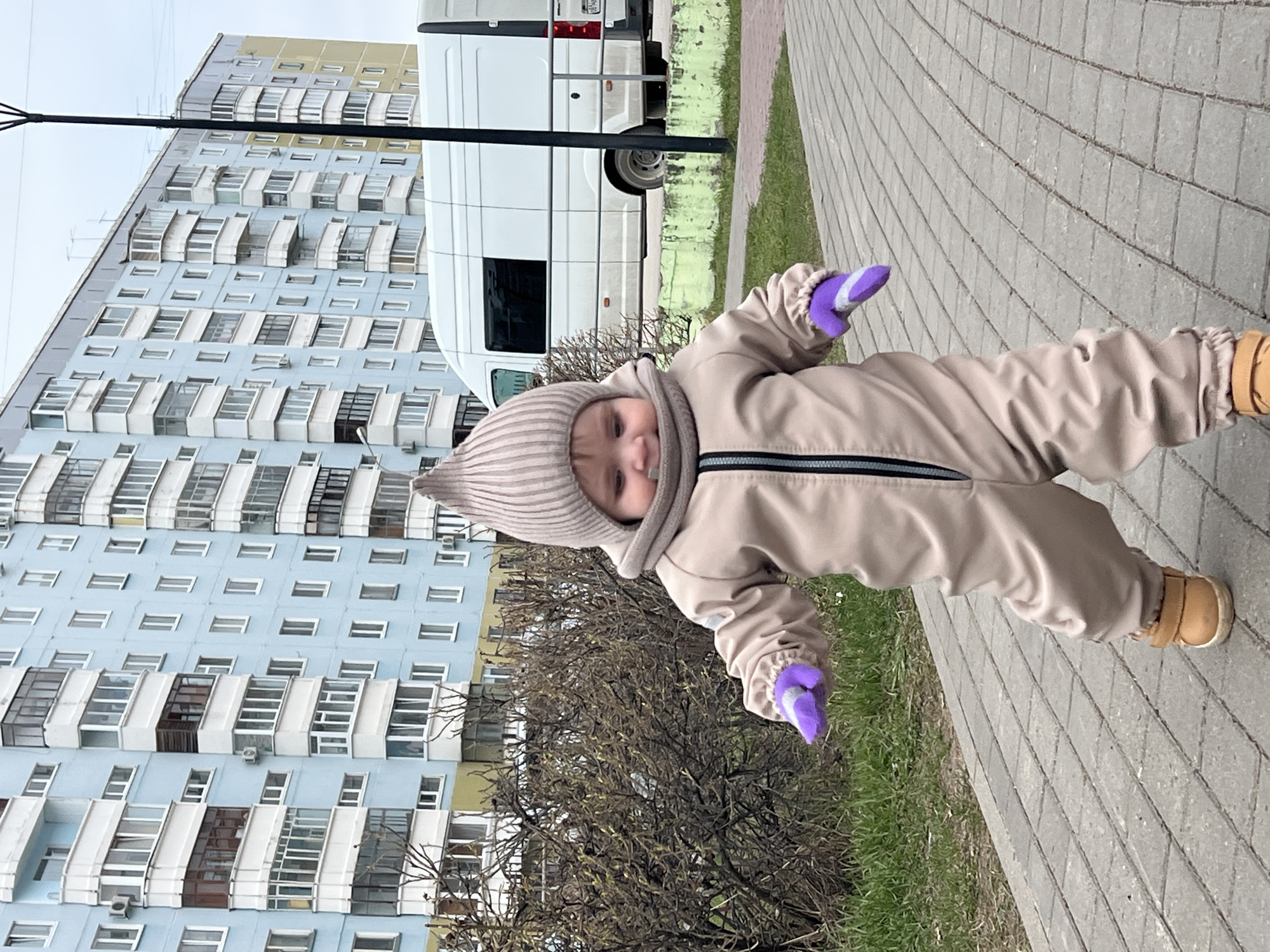 An outdoor photograph, presented at a tilt, features a young child, likely a toddler, standing on a grey paved sidewalk. The child wears a full-body beige jumpsuit with a dark grey zipper, a light beige and grey striped knitted hat, and purple mittens on both hands. Light yellow-brown shoes complete their outfit. The child looks towards the camera with a gentle smile, appearing content. Their pose suggests they are actively balancing or taking steps.
The immediate surroundings include bare deciduous bushes to the child's left and a strip of green grass running alongside the sidewalk. The background is dominated by a tall, multi-story apartment building with a light blue-grey facade. This building features numerous windows, many of which are part of enclosed balconies or loggias, some appearing white and others dark brown. Air conditioning units are visible on several balconies. A white commercial van or minibus is parked on the street in front of the building, with a partial license plate visible on its rear. A dark pole extends diagonally across the upper left section of the image.
The scene unfolds during daytime, under what appears to be an overcast sky, casting soft, diffused light. The child's warm attire indicates cool weather, suggesting early spring or late autumn. The architectural style of the residential building is characteristic of urban areas in Perm, Russia, aligning with the provided location context. No other legible text is visible in the frame.
