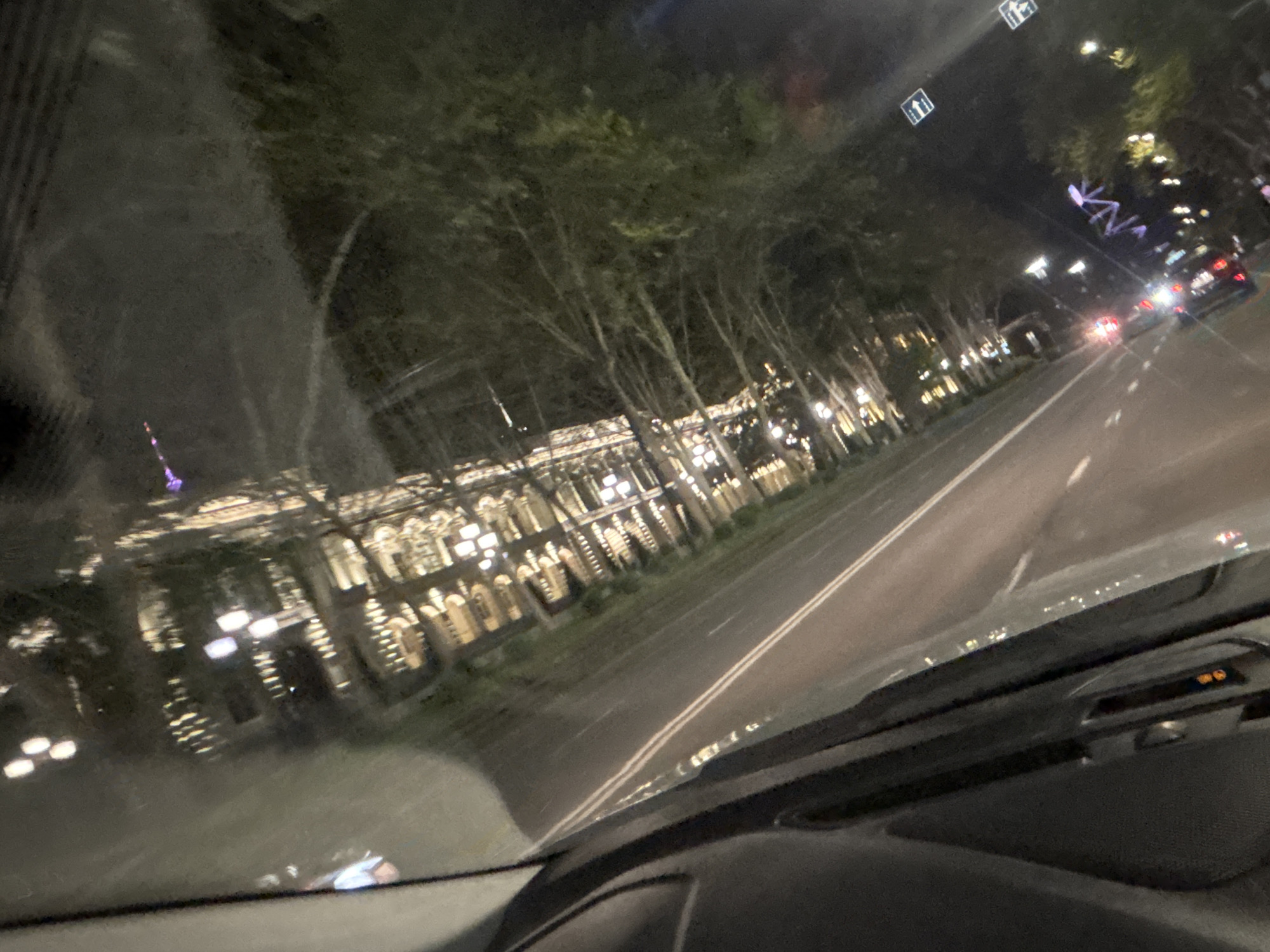 A night-time photograph taken from inside a vehicle, looking out through the windshield. The view is tilted diagonally to the right, showing a city street lined with trees and illuminated buildings.
On the left, a long, ornate building stands prominently, its facade brightly lit with warm white lights that emphasize numerous arched windows or architectural details. Trees, some appearing bare or with sparse foliage, line the street between the viewer and the building, their branches reaching upwards.
The two-lane road in the foreground and center has double white lines marking its middle. Reflections of lights are visible on the road surface, suggesting it might be wet or glossy. Further down the road, red tail lights of other vehicles are visible, indicating traffic or parked cars.
The windshield itself shows significant reflections, including portions of the car's interior (like the dashboard at the bottom) and distorted light sources, creating a layered visual effect. In the upper right quadrant, two square road signs, likely pedestrian crossing indicators, are visible with white stick figures on a blue background. A small orange indicator light can be seen on the dashboard in the bottom right, possibly related to airbag status, though specific text is not legible. The overall impression is an urban scene at night, captured from a moving or stationary car.