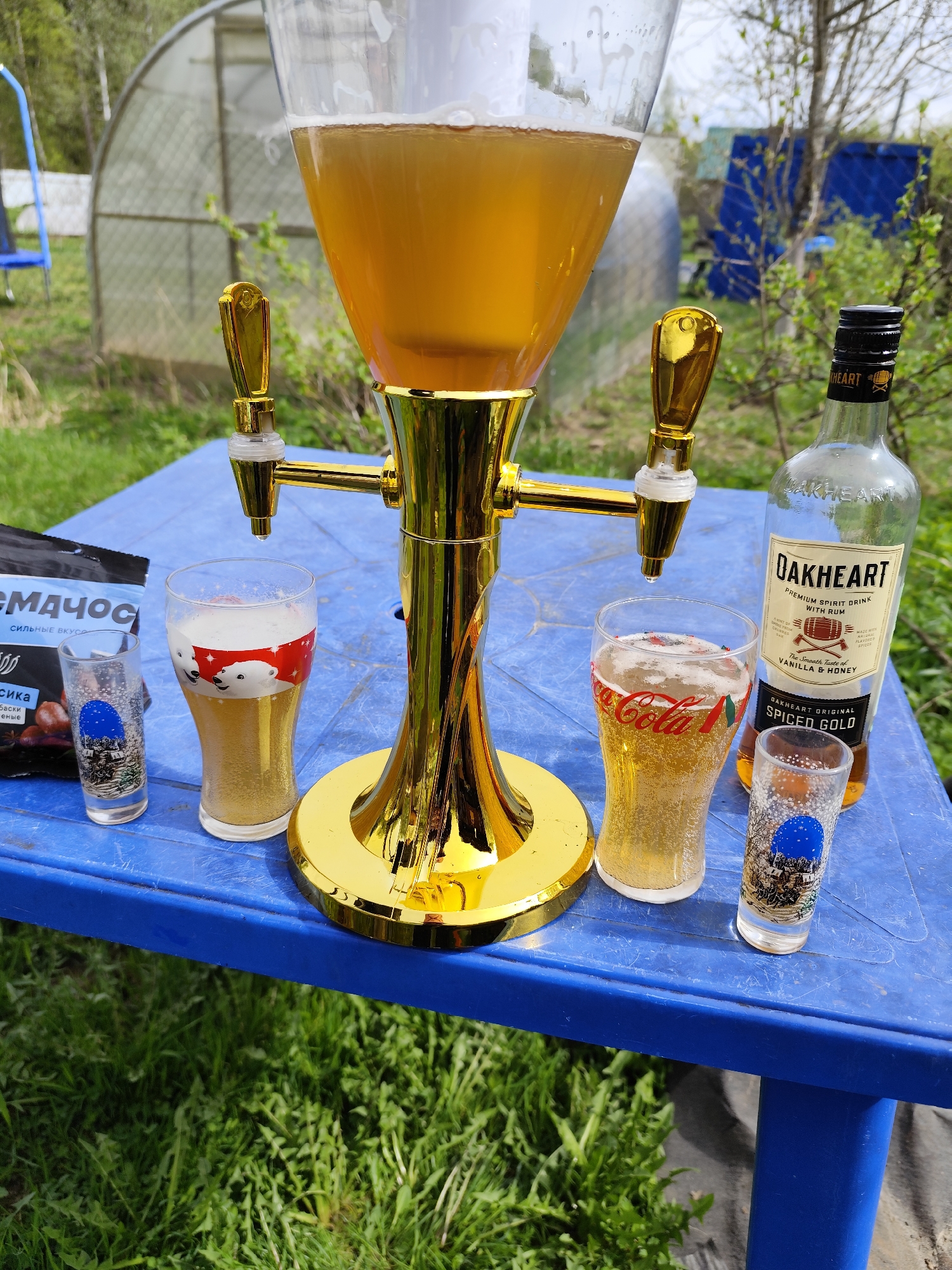 The image shows a blue plastic table outdoors, possibly in a garden or yard, with various items placed on it. A large, clear glass dispenser filled with a light-colored beverage with a gold-colored stand and two spouts dominates the center of the table. To the left of the dispenser is a bag of "СМАЧОС" snacks and a small shot glass with a blue image on it. Next to the snacks is a taller glass with a red and white polar bear design, filled with a light-colored liquid. To the right of the dispenser, there's a bottle of OAKHEART spiced gold rum and a Coca-Cola glass filled with liquid. Another small shot glass with a blue image is placed next to the rum. The background includes green grass, trees, and a greenhouse structure, suggesting the image was taken in a rural or suburban setting, potentially near the city of Афанасьево, Russia. The scene appears to be set up for relaxation and enjoyment of drinks and snacks outdoors.
