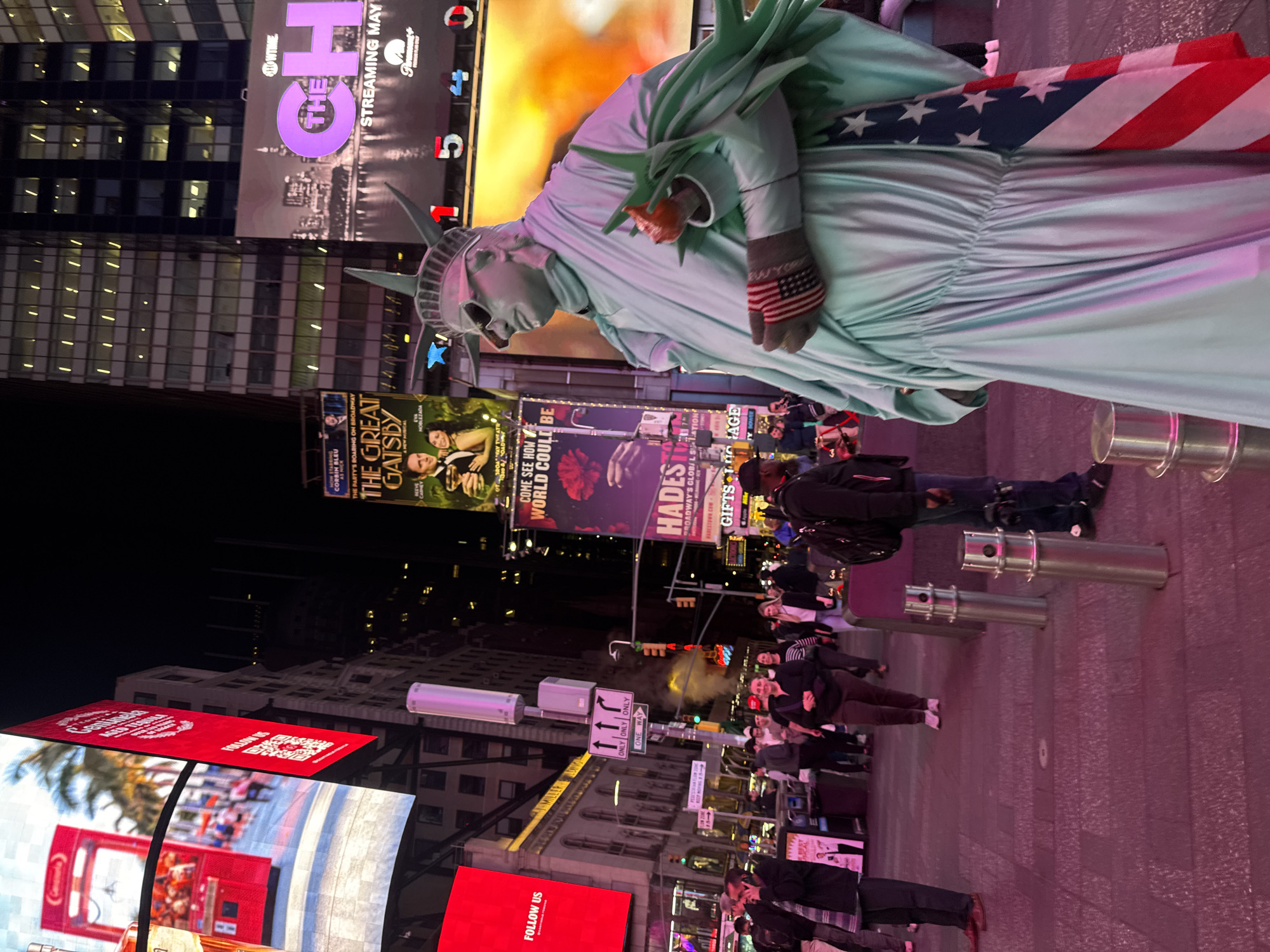 The image shows a human statue of the Statue of Liberty in Times Square, New York City. The statue is wearing a costume resembling the Statue of Liberty, with a US flag draped over its shoulder. The background consists of illuminated billboards and street signs, indicating that it is night. People are walking around, and there are advertisements for "The Great Gatsby" and "Hades" shows, along with a Showtime promotion.