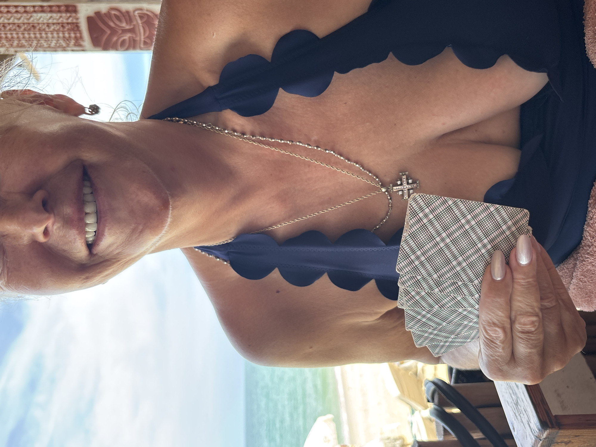 A woman with short, blonde hair is smiling and holding a stack of playing cards. She is wearing a dark blue scalloped swimsuit top and a silver necklace with a cross pendant. The background shows a beach and ocean, indicating an outdoor location. The photo was taken in Baan Hua Thanon, Thailand.