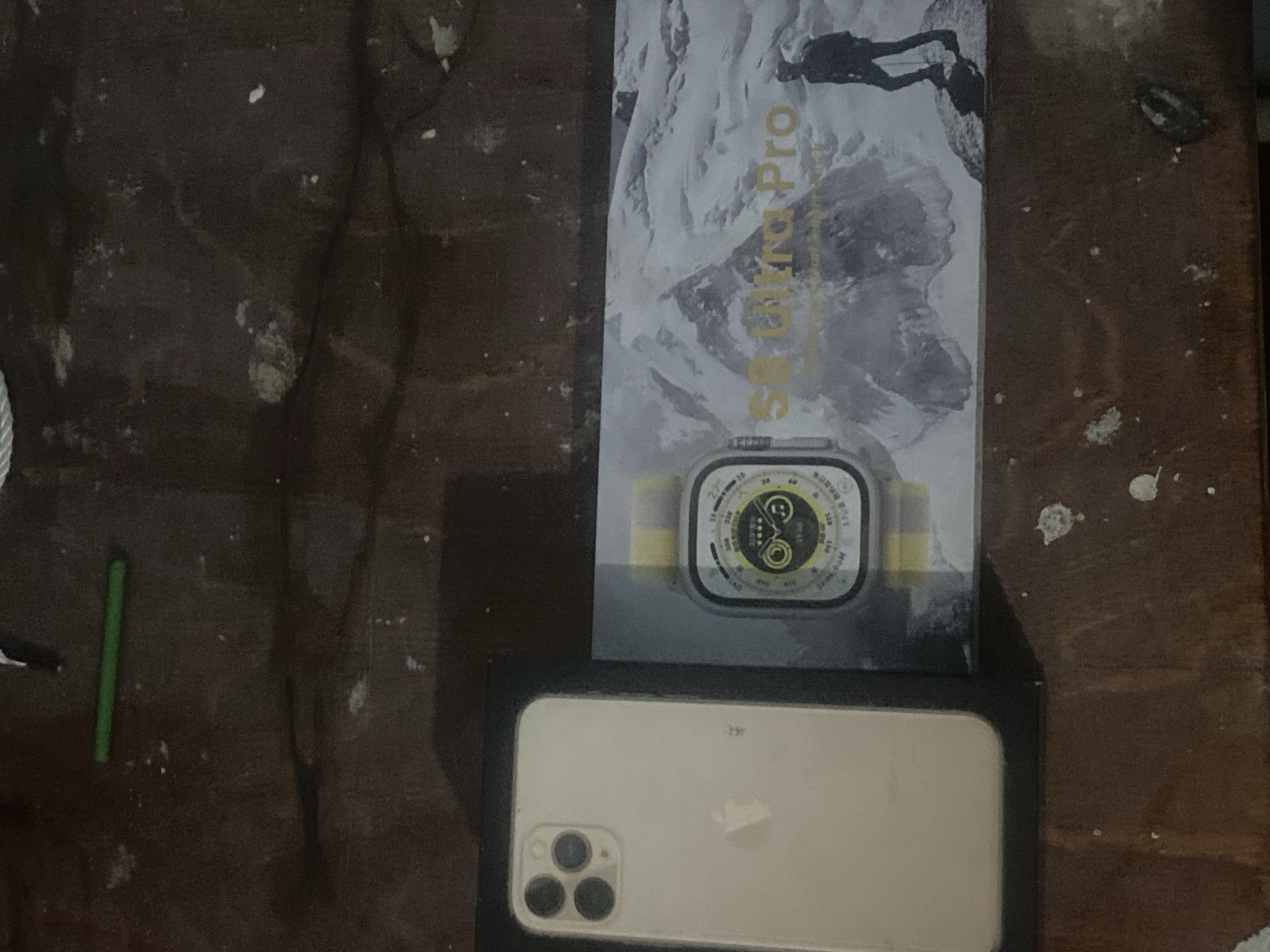 The image shows a smartphone and a smartwatch box on a dark, wooden surface. The smartphone is a light-colored iPhone with three camera lenses on the back. The smartwatch box is rectangular and features an image of a mountainous landscape with a person hiking. The text on the box reads "S8 Ultra Pro". The scene is likely indoors, as indicated by the artificial lighting and the presence of a green object that resembles a pen or a pencil lying on the surface in the top left corner. The surface appears somewhat dirty with white specks scattered across it. No specific location in Dusti, Tajikistan can be determined from the image itself.