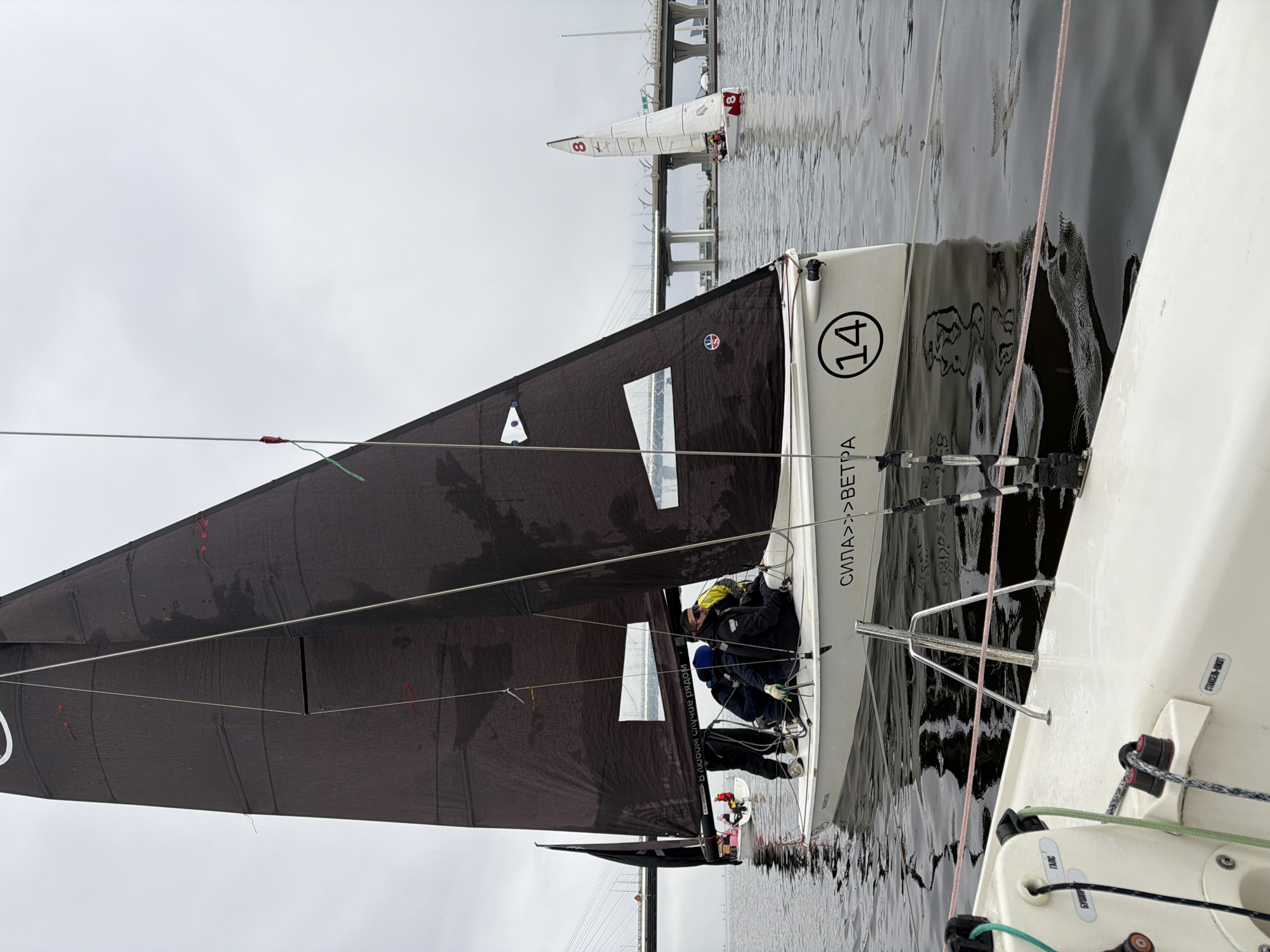 The image shows two sailboats on the water, potentially participating in a race. The sailboat in the foreground is marked with the number "14" and Russian text including "СИЛА>>>>ВЕТРА" and "В любом случае рядом." The sails of this boat are a dark color. Two people are visible on board, wearing what appears to be sailing gear. Another sailboat, marked with the number "8", is visible in the background. A bridge spans the waterway, suggesting an urban setting. Based on the Russian text and the type of bridge structure, it's highly probable that the location is Saint Petersburg, Russia. The sky is overcast, indicating potentially dull or rainy weather.