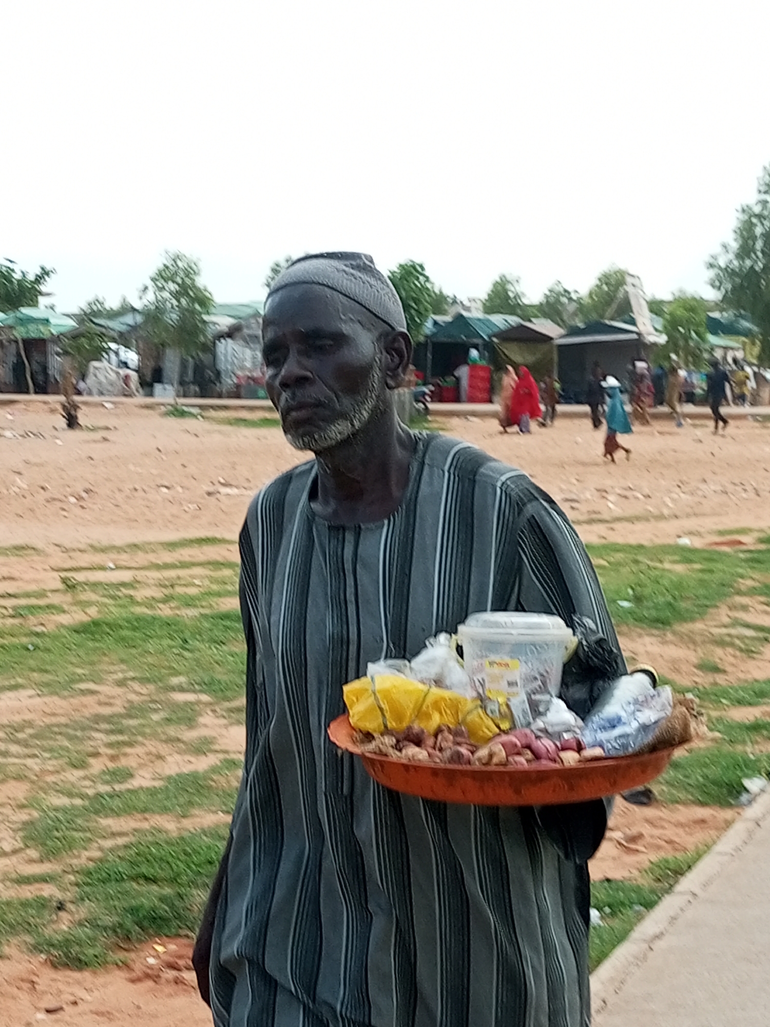 An elderly African man, wearing a grey and white striped traditional garment and a matching skullcap, stands in the foreground looking slightly to his right. He has a grey beard and his skin is dark. He is holding a shallow, orange tray filled with various items. These include what appear to be onions, small red and yellow objects wrapped in plastic bags, and a clear plastic container with a lid. In the background, there are several makeshift structures that resemble market stalls or temporary shelters, some with green tarpaulin roofs. People, some dressed in colorful clothing, can be seen in the distance, moving around the area. The ground is a mix of sandy soil and sparse green grass. The sky is overcast and pale. The scene appears to be an outdoor market or community gathering in Gombe, Nigeria.