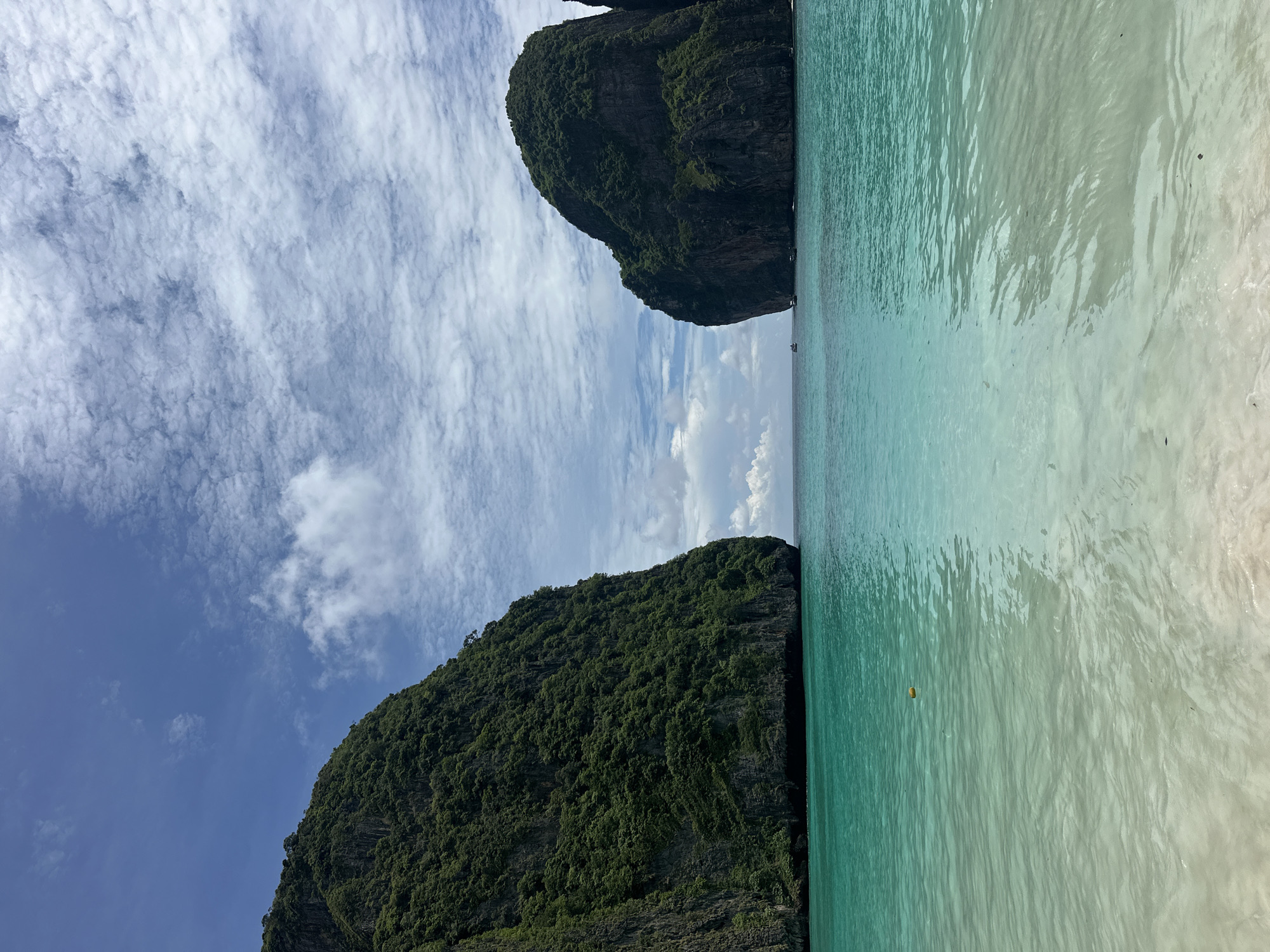 This image depicts a scenic view of Maya Bay, located in the Phi Phi Islands, near Ao Nang, Thailand. The bay is characterized by towering limestone cliffs covered in lush green vegetation on either side, creating a dramatic entrance. The water is exceptionally clear and ranges in color from a vibrant turquoise near the shore to a deeper blue further out. The shallow areas near the beach reveal a sandy bottom. The sky is partially cloudy, with white, wispy clouds scattered across a bright blue expanse. In the distance, a few small boats are visible on the water, suggesting the presence of other visitors or tours. A yellow buoy floats in the water closer to the shore. The overall atmosphere is one of tropical tranquility and natural beauty.