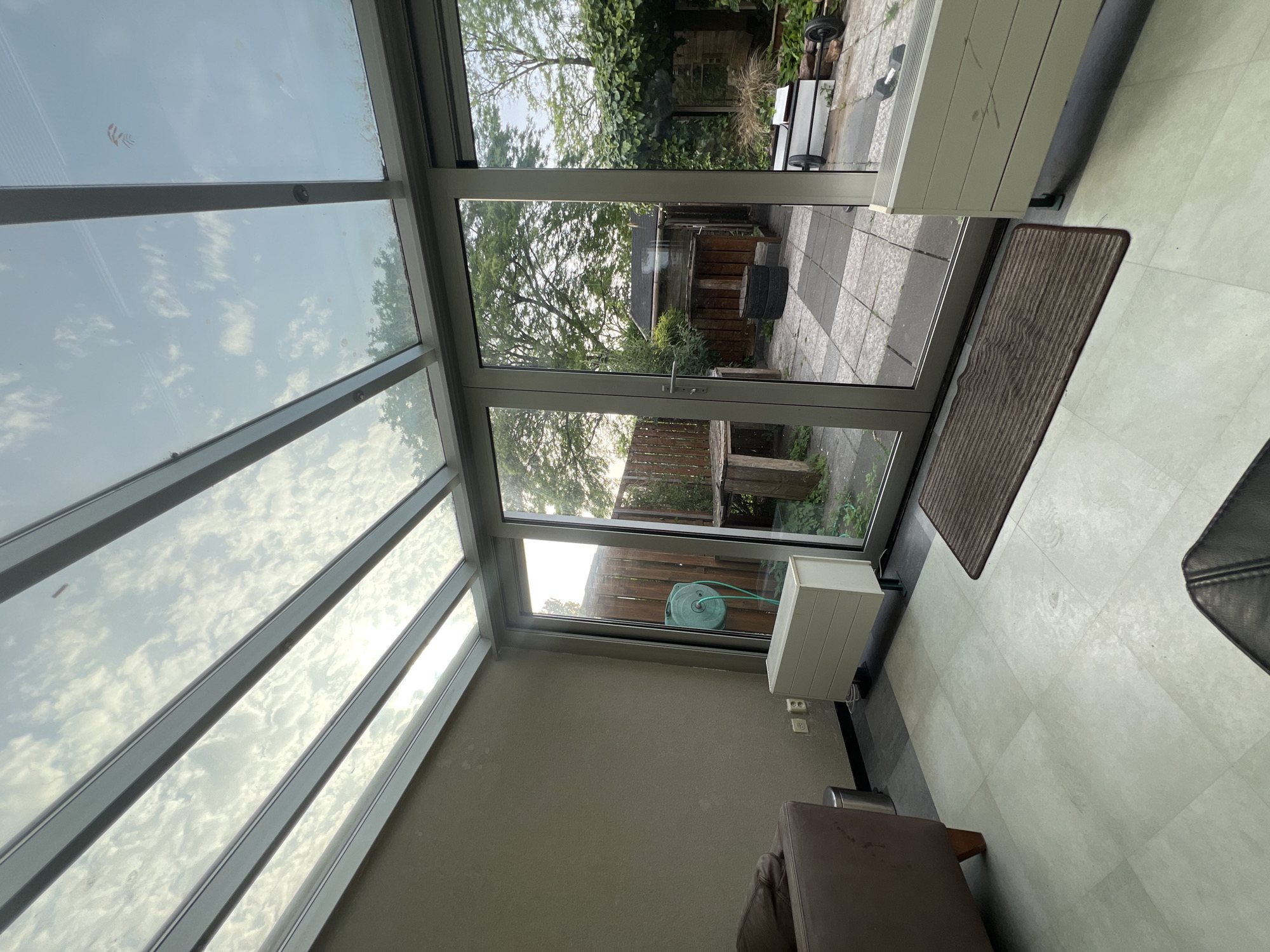 The image depicts the interior of a conservatory or sunroom. Light enters through the angled glass roof, reflecting the sky and clouds. The floor is tiled in a light color. Outside the large glass doors is a garden area with plants and a fence. A coiled blue garden hose is mounted on the wall inside the conservatory. Two white rectangular objects, possibly air conditioners or heaters, are visible. A brown rug lies near the entrance. A portion of furniture is visible, suggesting the space is used for seating. Based on the request, it is inferred that the location of the image is Oud-Beijerland, Netherlands.