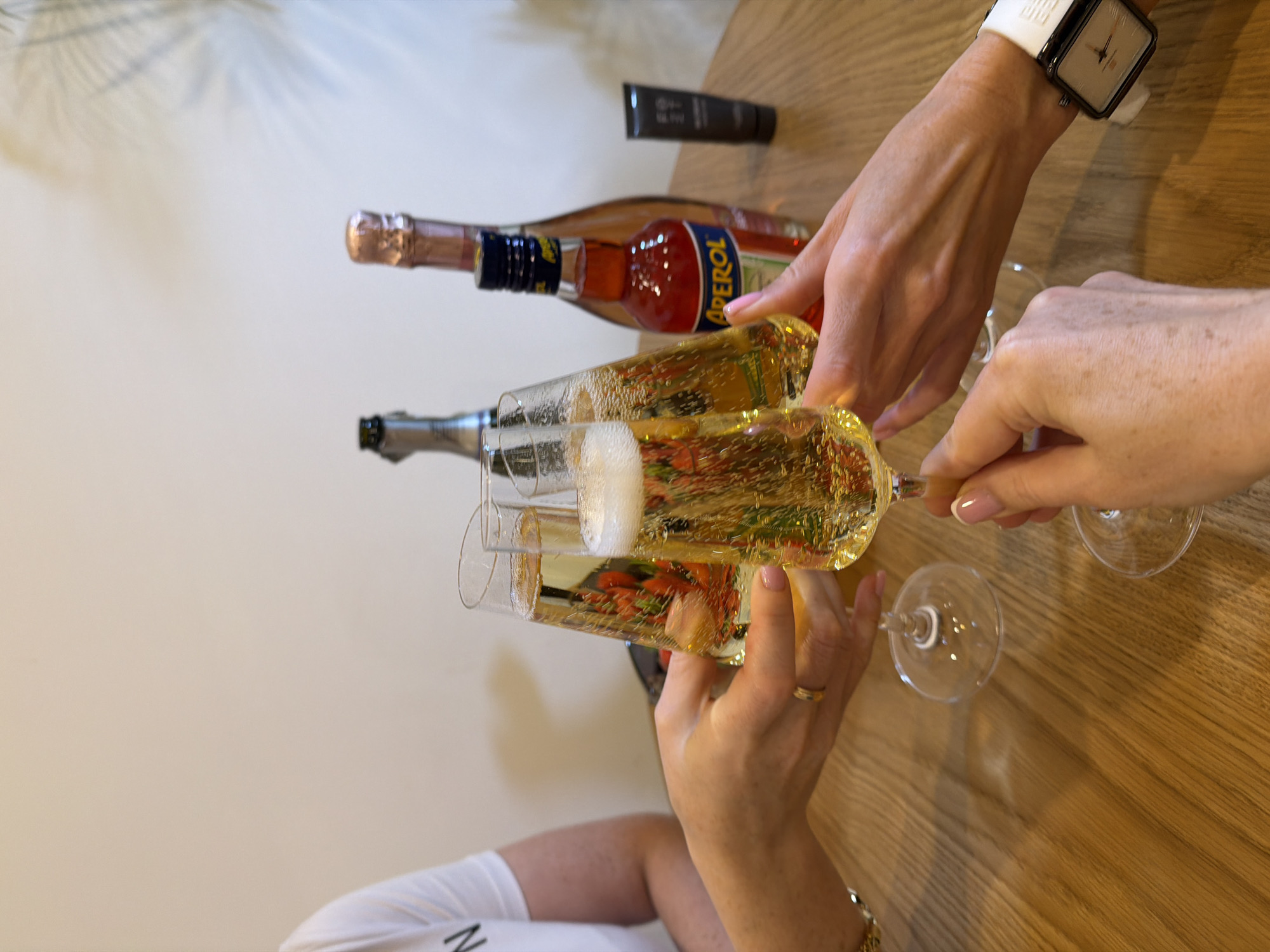 On a light wooden table, three wine glasses filled with champagne are being held in hands toasting, with two bottles of wine and a small tube of product in the background. One bottle is light red with a prominent "APEROL" label, and the other is silver. The hand holding one of the glasses has a gold ring and a gold bracelet. The hand visible on the upper right wears a white wristwatch with a dark frame. A shadow from a plant or tree is cast on the wall in the upper left corner. The location is not identifiable as Khimki, Russia, from the provided image.