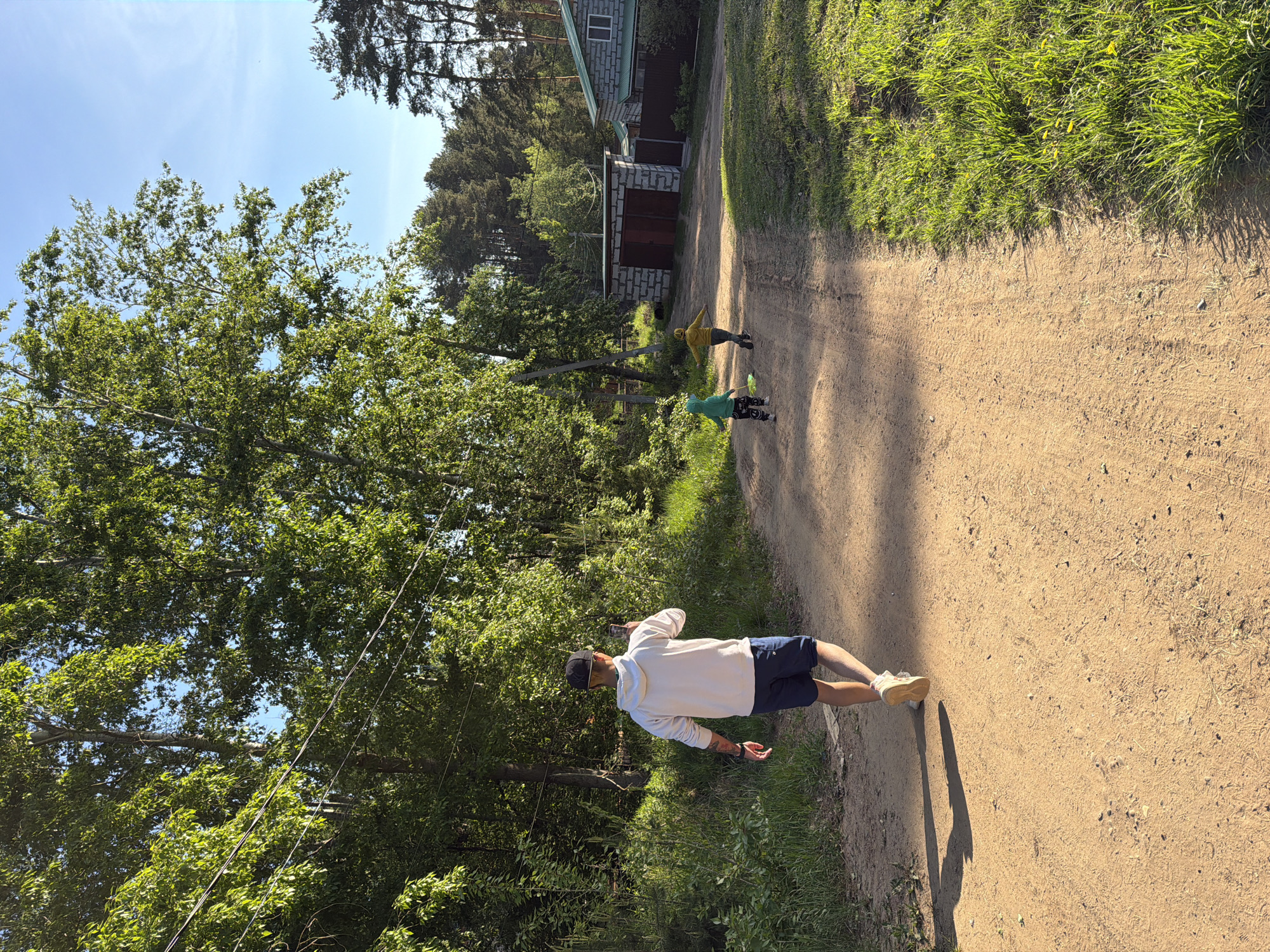 The image shows a dirt road in a wooded area, likely on the outskirts of a town. The location is the city of Chaikovsky, Russia. The road is flanked by trees and grassy areas. In the middle ground, there are two children walking or running away from the camera. One is wearing a yellow jacket and the other a green one. In the foreground, a man is walking in the same direction as the children, but on the edge of the road. He is wearing a white shirt, dark shorts, a black cap, and light-colored sneakers. He seems to be holding a phone and looking at it. A building is visible in the background, beyond the children. The sky is blue and the weather seems sunny.