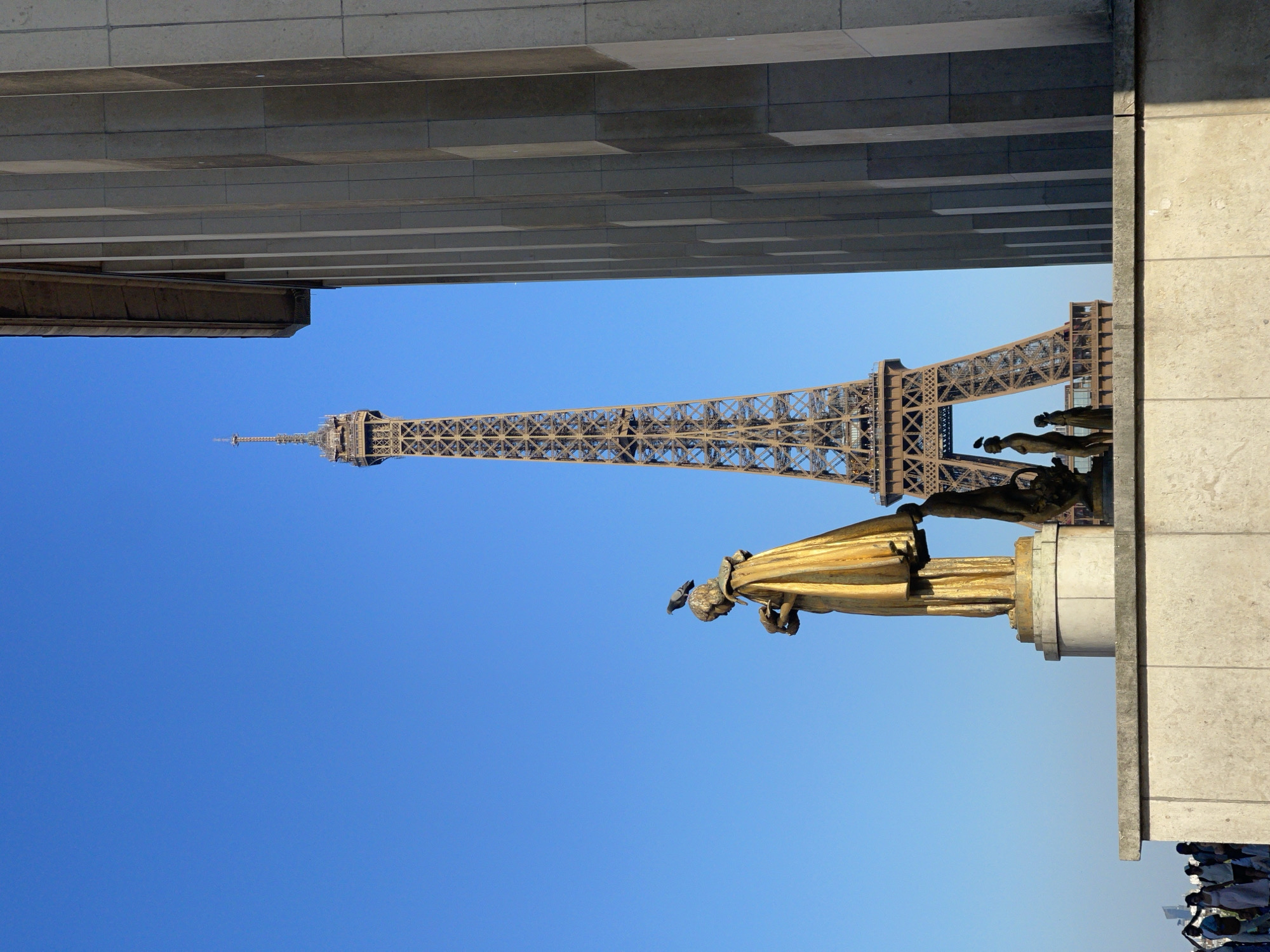 This is a photograph of the Eiffel Tower in Paris, France, taken from an angle that includes a large stone building with vertical columns on the right side of the frame. In the foreground, a golden statue of a woman holding what appears to be a bird is prominently displayed. A pigeon is perched on the head of the statue. To the right of the main statue, there are smaller bronze statues, one of which is a woman holding a basket. In the background, the Eiffel Tower is visible through a gap between the foreground statue and the stone building. The sky is clear and blue. At the bottom left of the image, a group of people are walking along a ledge, some of them silhouetted against the sky. The scene is captured during daylight.