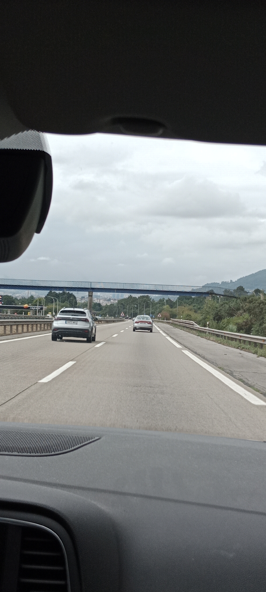 The image is a photograph taken from inside a vehicle, looking forward on a highway. The highway has multiple lanes, separated by white lines. In the distance, there is a blue pedestrian bridge spanning the highway. On either side of the highway, there are trees and hills, with a city visible in the background. Two cars are visible on the highway; one silver SUV in the left lane and a silver sedan in the right lane. The sky is overcast with clouds. The foreground shows the dashboard of the vehicle, including a speaker grill and part of a vent. This scene is consistent with the A-66 highway near Llanera, Spain, given the distinctive blue pedestrian bridge often seen on this route and the surrounding landscape.