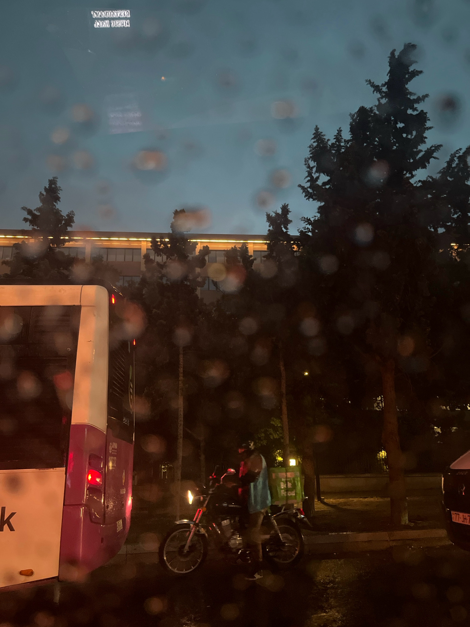 In an urban setting at dusk, a scene unfolds through a rain-streaked window. On the left, the rear of a white and purple bus is visible, its brake lights illuminated. To the right of the bus, a person wearing a helmet and a light blue garment stands beside a motorcycle. The motorcycle has a green food delivery box attached to its rear. The surrounding environment includes trees and a building with illuminated windows in the background. The ground appears wet, suggesting recent rain. The sky is a deep blue, indicative of twilight.