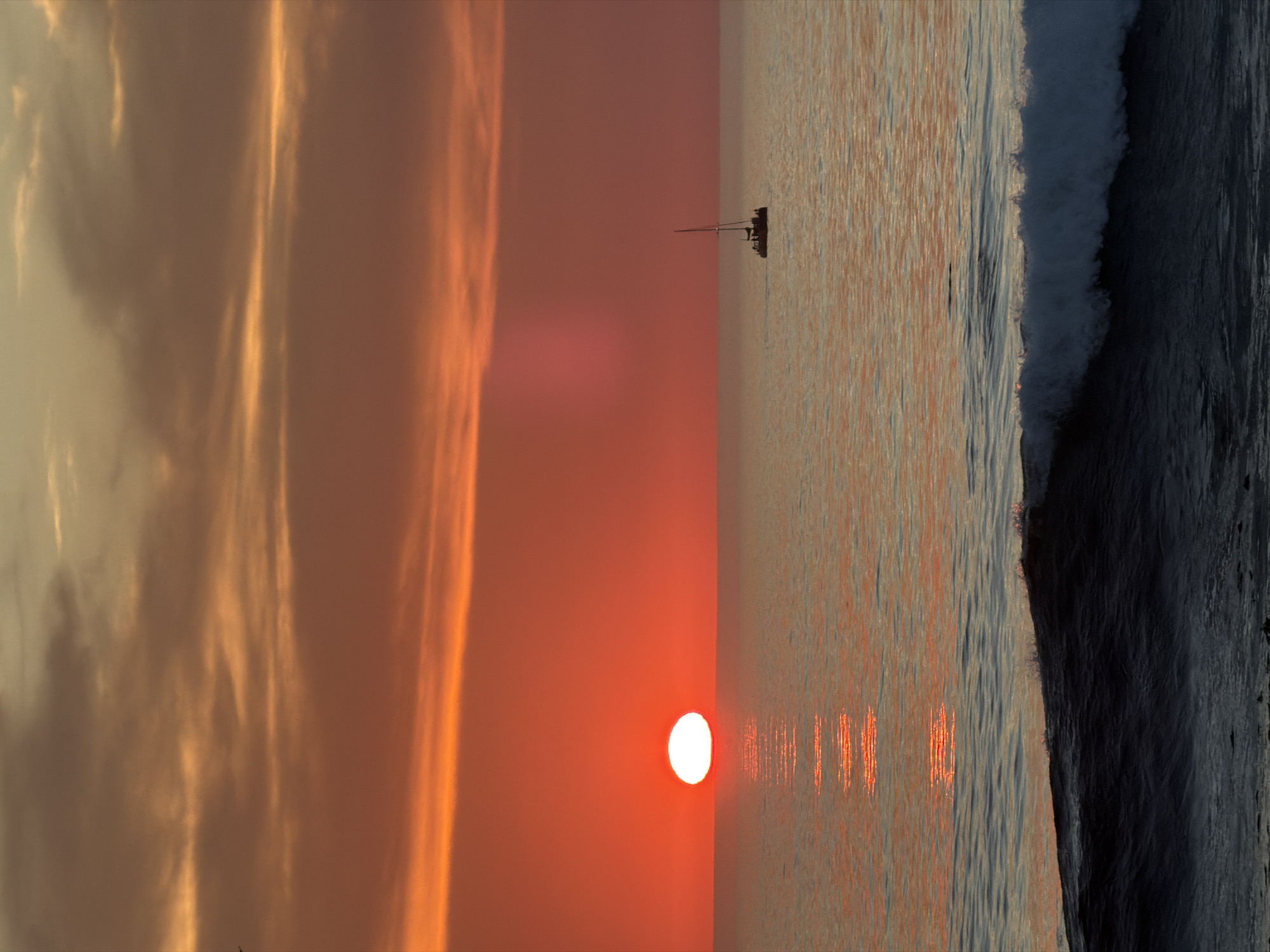 The image depicts a sunset over the ocean, likely viewed from the coast of Cape Town, South Africa. The sun, a bright orange disc, is positioned low on the horizon, casting a warm glow across the sky and water. The sky is filled with dramatic, orange and pink clouds, suggesting a vibrant sunset. In the middle distance, a small sailboat with a single mast is visible, its silhouette stark against the colorful sky. The ocean in the foreground features gentle waves and a dark, foamy crest, indicating the approach of a breaking wave. The overall scene evokes a sense of tranquility and natural beauty, characteristic of coastal sunsets in the region.