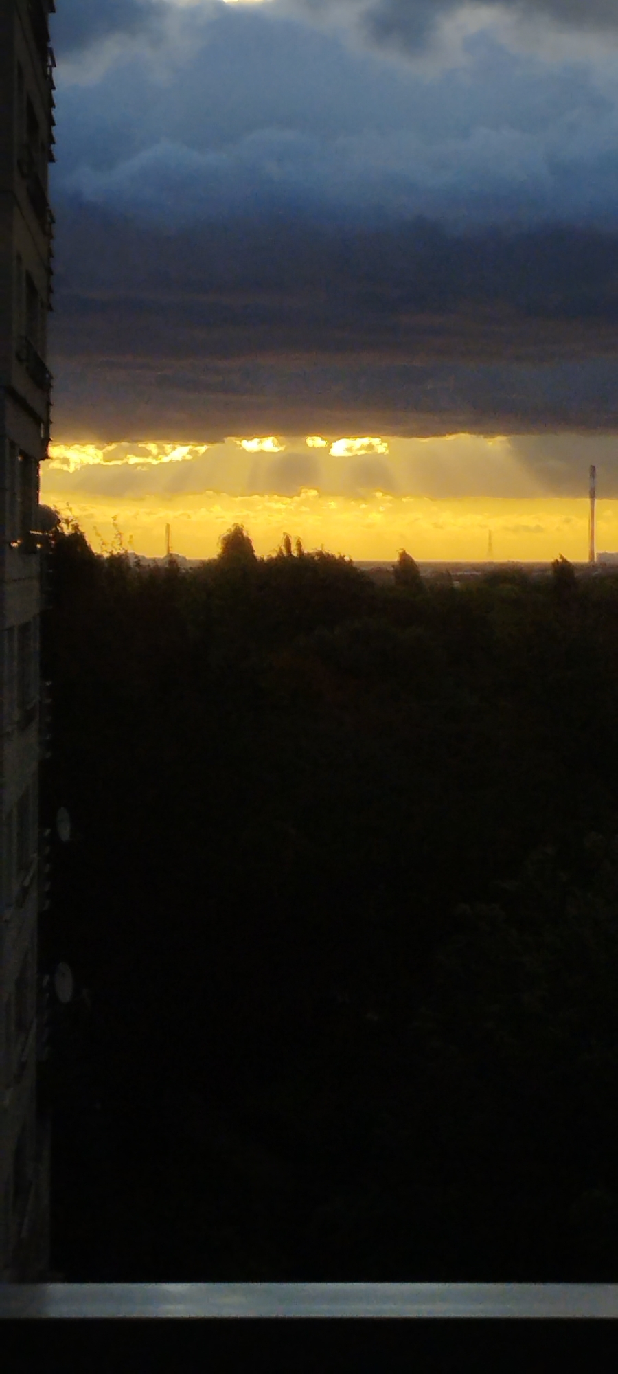 This is a photograph of a sunset viewed from a window in Warsaw, Poland. The sky is filled with dramatic clouds, with a bright band of sunlight peeking through near the horizon. The sun's rays are visible, creating a dramatic effect. In the foreground, the dark silhouette of trees can be seen, with a tall, industrial-looking tower visible in the distance. On the left side of the image, the corner of a building is visible, also silhouetted against the bright sky. The scene is dark and moody, with the exception of the bright band of light from the setting sun.