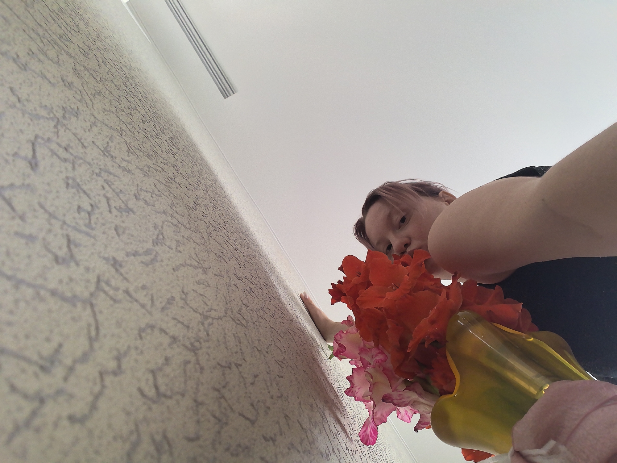 The image shows a person holding a bouquet of flowers against a textured wall. The person has light-colored hair with pinkish tones. The bouquet contains vibrant red and pink flowers. A yellow vase is visible supporting the flowers. The background appears to be a light-colored ceiling with what looks like a vent or grille. The perspective is from a low angle, looking up at the person and the flowers. The environment seems to be an indoor setting. There is no information to suggest this image is located in Chaikovsky, Russia.