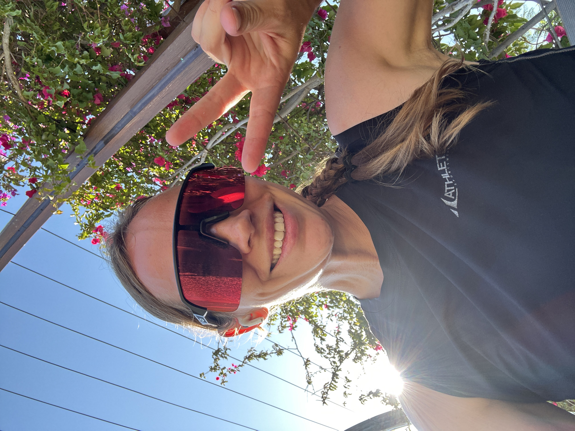 This vibrant selfie captures a smiling woman outdoors in Fethiye, Turkey, presented in a counter-clockwise rotated orientation. The woman is positioned on her right side, looking upwards and towards the viewer. She is seen from the chest up, wearing large, reflective red-tinted sports sunglasses that obscure her eyes. Her light brown hair is styled in a braid, and she wears a black t-shirt with a white logo partially visible, reading "ATHLETE SPORTSWEAR". Her face expresses happiness through a wide, open-mouthed smile, showing her teeth. Her right hand is raised above her head, making a peace sign (V-sign) gesture with her index and middle fingers.
The background primarily features lush green foliage intertwined with vibrant pink bougainvillea flowers and wooden pergola beams, filling the upper part of the rotated frame. A clear, bright blue sky occupies the lower-left section. A strong lens flare from the sun bursts from the lower-right corner of the image, indicating bright, sunny weather, likely either morning or late afternoon. The overall mood is cheerful and positive, suggesting a pleasant outdoor moment or active leisure time in a sunny, scenic location.