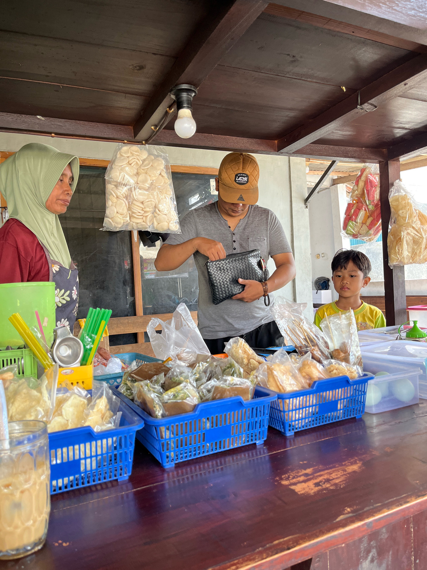 In Banguntapan, Indonesia, a modest outdoor food stall operates under a wooden roof. A woman, wearing a light green hijab and a maroon shirt with a floral apron, stands behind a counter filled with various snacks. To her right, a man in a grey t-shirt and a brown baseball cap examines a black zippered pouch. A young boy in a yellow t-shirt sits to the man's right, looking forward. The counter displays blue plastic baskets overflowing with packaged snacks, including what appears to be crackers and fried goods. To the left, a green plastic container holds yellow and green straws, and a glass of iced coffee sits nearby. Bags of light-colored crackers hang from the ceiling.