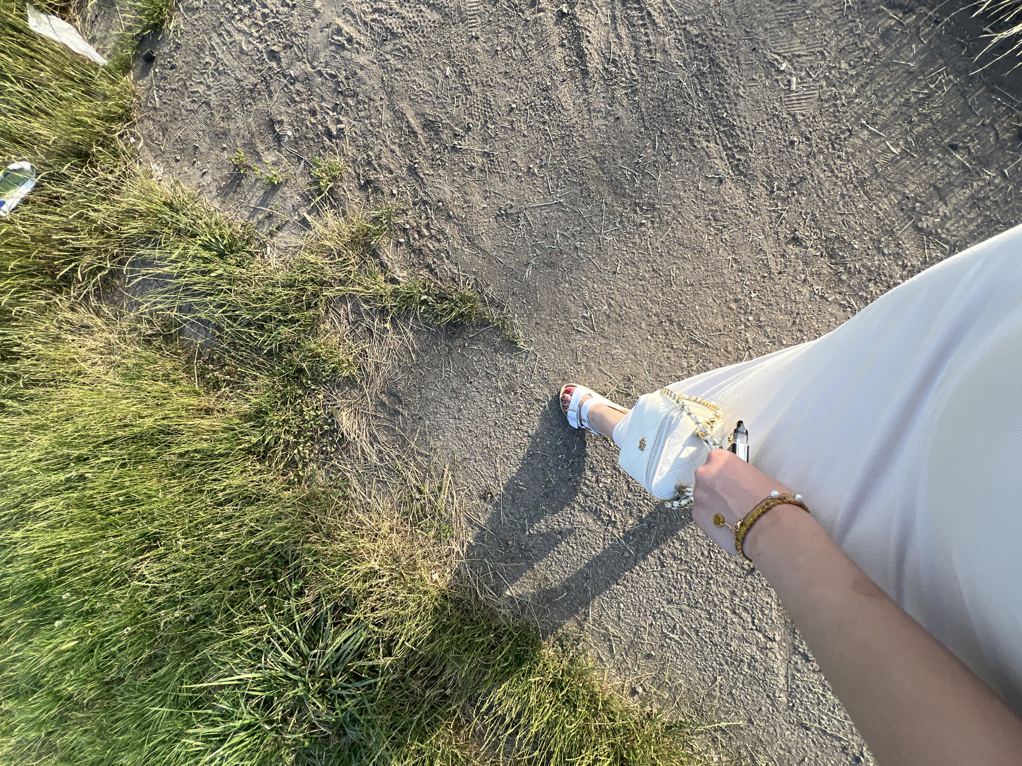 A first-person perspective shot shows a person's legs and arm as they walk on a dirt path next to a grassy area. The person is wearing a light-colored dress, white sandals with red accents, and a white quilted handbag with a gold chain. They are also wearing a gold bracelet with charms. The path has tire tracks and footprints visible. The grassy area is lush and green. The lighting suggests it is late afternoon or early evening. The scene is likely outdoors in a park or a rural area.