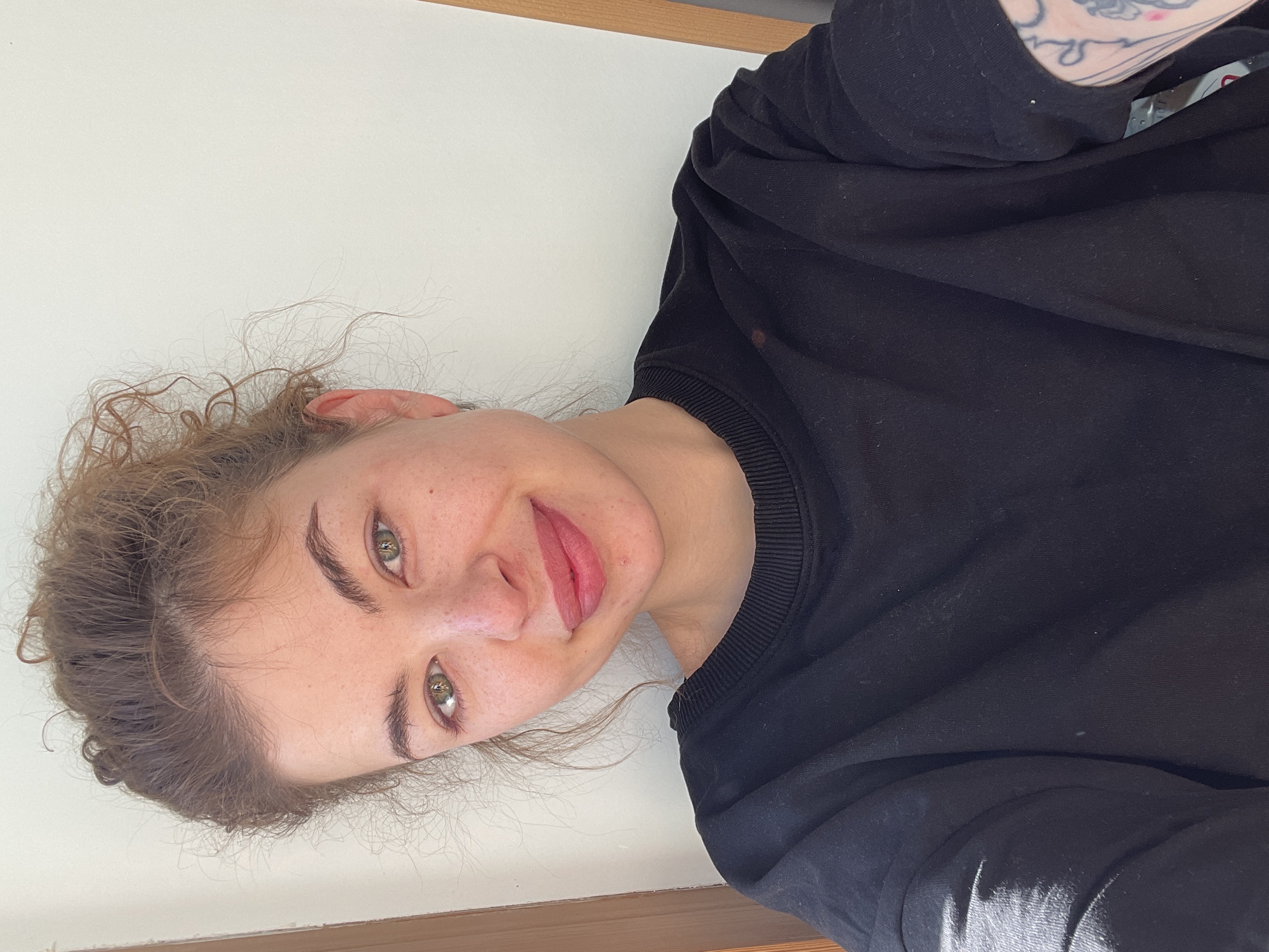 A woman with fair skin, green eyes, and light brown curly hair is pictured indoors against a white wall with a wooden trim along the base. She is wearing a black sweatshirt. Part of a tattoo is visible on her upper arm. The image appears to be taken from a slightly elevated perspective. There is no evidence of the location being Tbilisi, Georgia.