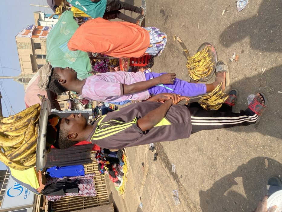 Two men are sitting beside each other, each holding a tray of bananas. One tray is on his head, while the other is on the ground. The men are dressed in casual attire, t-shirts, and trousers. They are near a clothing stall with colorful clothes. The background includes a building and other people. The setting appears to be a busy street market or outdoor vending area.