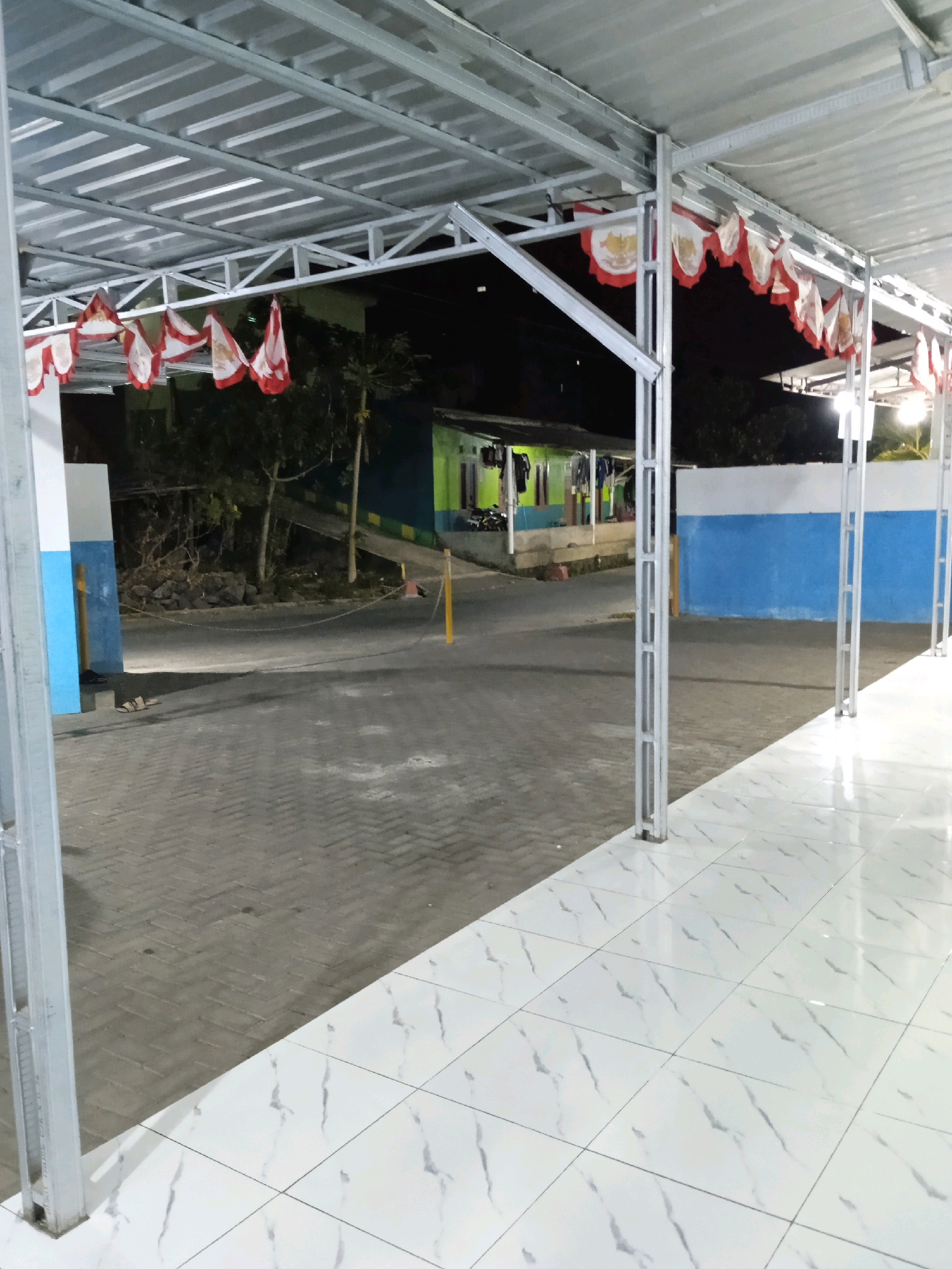The image captures an outdoor area under a metal canopy at night, with a paved walkway in the foreground and a road or courtyard beyond. The walkway is covered by a white tiled floor with a marble-like pattern, and is supported by thin metal posts. The canopy itself is constructed from corrugated metal sheeting and a metal frame. Decorative triangular flags, red and white, are strung along the edge of the canopy.
In the background, a low-rise building with green and blue painted walls is visible. Clothes are hanging on a line outside the building. The ground behind the canopy is paved with grey interlocking bricks. To the left, a light blue and white painted pillar stands next to the walkway. A pair of flip-flops are visible on the grey brick ground near the pillar. A yellow traffic cone and a rope barrier are present on the road beyond the canopy. Trees and foliage are sparsely visible behind the building, suggesting a semi-urban or rural setting. The overall lighting indicates it is nighttime, likely illuminated by artificial lights. The location is identified as Wringinputih, Indonesia.