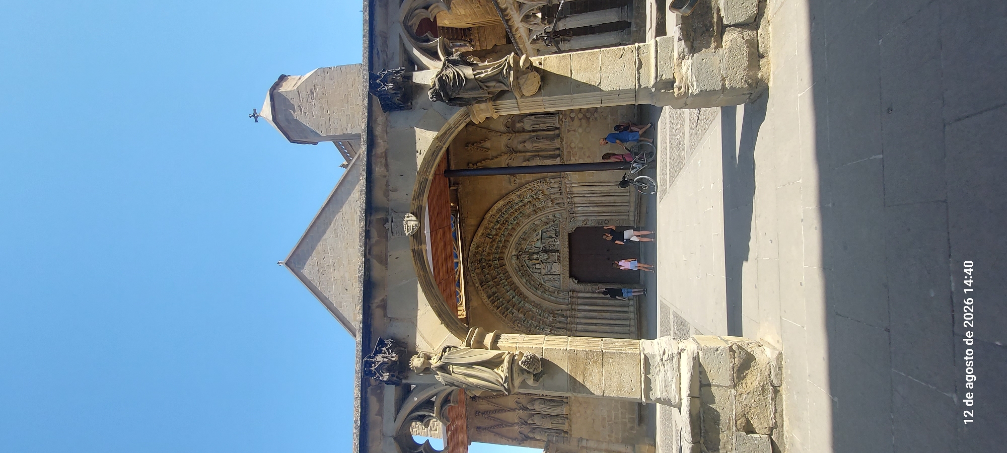 The image depicts the entrance to the Palace of the Kings of Navarre in Olite, Spain, on August 12, 2026, at 14:40. The scene is a wide-angle shot of the ornate stone entrance of a historical building, likely a cathedral or palace, under a clear blue sky.
In the foreground, the stone paving stones of a courtyard are visible, with a prominent shadow cast from the right side, suggesting it is midday. Two large, weathered stone pillars frame the entrance. These pillars are decorated with intricate carvings and topped with statues. The left pillar features a statue of a crowned figure in a draped robe, with hands clasped in prayer. The right pillar also has a statue, partially obscured by shadow, appearing to be a draped figure as well.
The entrance itself is a grand, arched gateway. The archway is richly decorated with a tympanum filled with detailed relief sculptures depicting a religious or historical scene. The main doors are large and dark, centrally positioned within the arch.
Several people are present, appearing to be tourists. On the left side, near the entrance, two children and an adult are standing, looking towards the building. Further inside the archway, a man in a dark shirt and shorts is pointing upwards. On the right side, near the entrance, a group of three people are gathered, one of whom is a man wearing a blue shirt and shorts, and another a woman in a pink dress. A bicycle with a rack is parked beside them.
The architecture is characteristic of Gothic or Romanesque styles, with elaborate stonework, pointed arches, and decorative elements. The overall impression is of a historical and culturally significant site on a bright, sunny day.