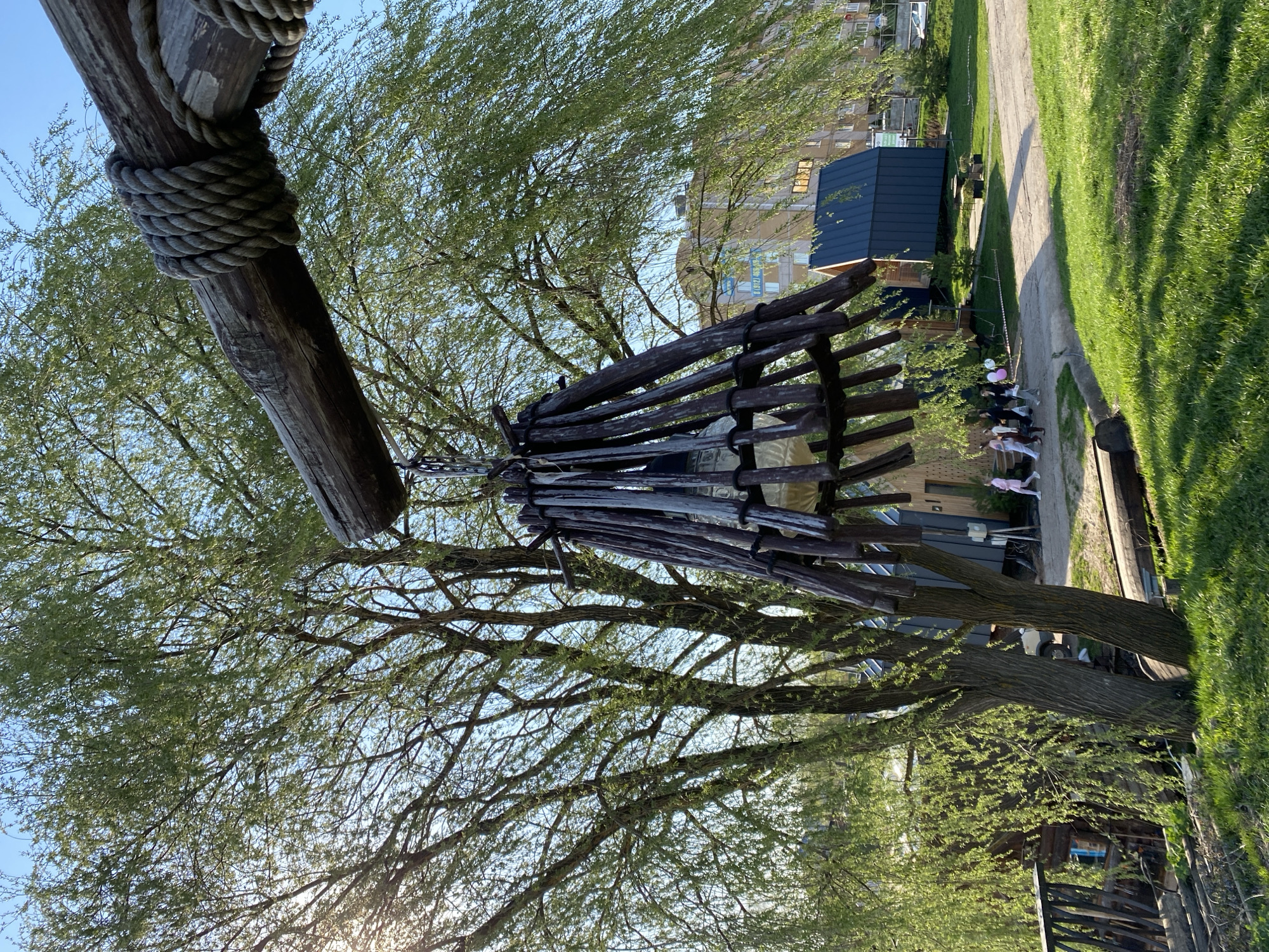 The image depicts a wooden structure, possibly a swing or art installation, in a park-like setting, potentially in Votkinsk, Russia. The structure consists of a thick wooden beam suspended by rope and a frame of dark wooden planks hanging below. A large tree with green leaves fills the background, with branches partially obscuring the wooden frame. In the distance, buildings are visible, including a dark blue shed-like structure and an apartment building. A grassy area is present, and a paved path runs alongside it. Several people are seated on a bench, with some holding balloons. The lighting suggests it's daytime, with some sun glare in the lower left.