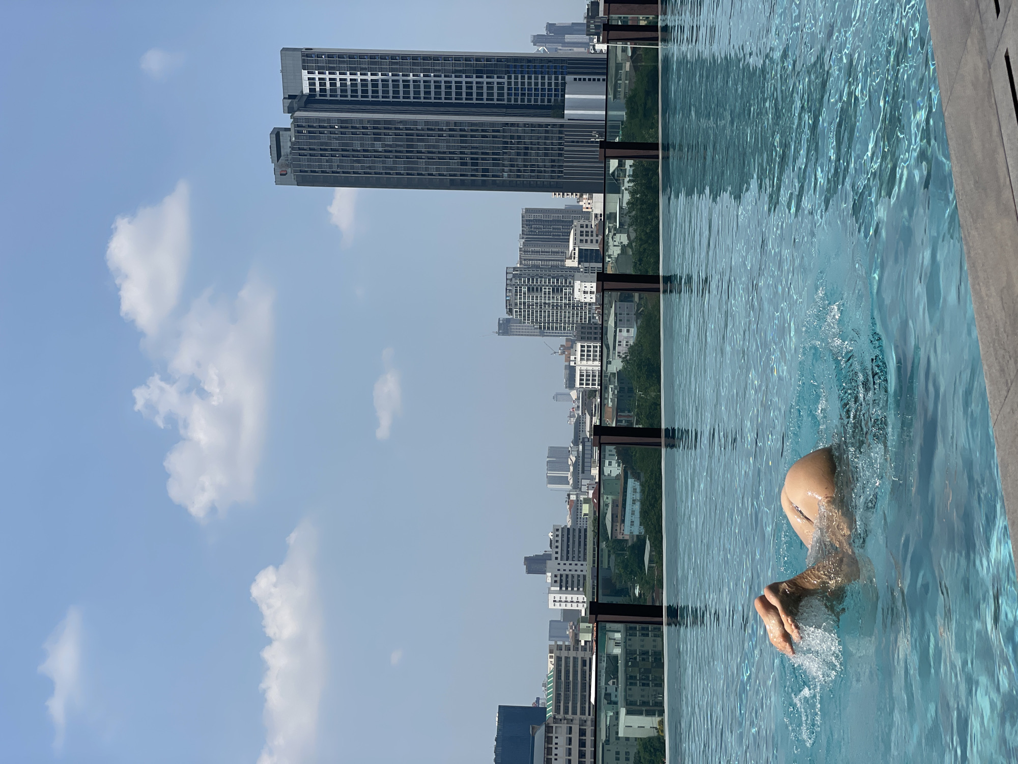 A vertical, eye-level shot shows a partial view of a person submerged in a rooftop infinity pool, set against the skyline of Bangkok, Thailand. The person's lower torso and legs are visible, submerged in the rippling, light blue water. The water reflects the cityscape, creating a mirrored effect. The pool's edge is a dark, horizontal line. The backdrop features numerous high-rise buildings, including a prominent, multi-story, grey skyscraper to the left. A mix of residential and commercial buildings are scattered across the landscape, partially obscured by trees. The sky is light blue, with scattered white clouds.
