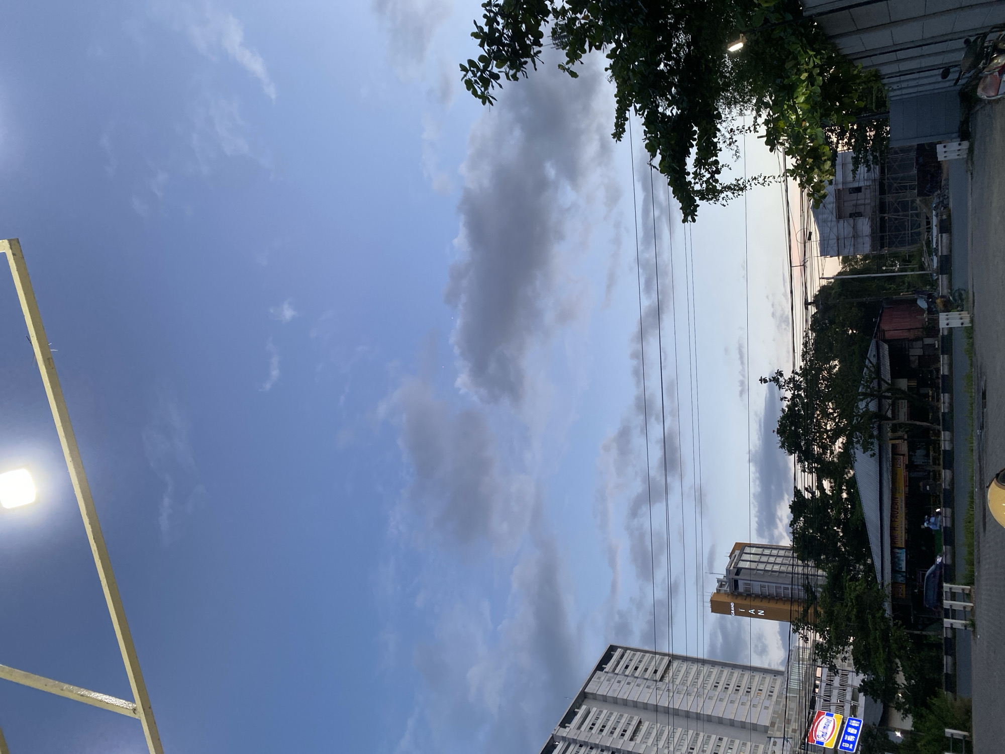 The image captures a dusk scene in Sungai Lakum, Indonesia, characterized by a vast expanse of cloudy blue sky above a low-rise urban landscape. In the foreground, a yellow metal structure with a bright light fixture is visible against the sky. Below, the cityscape unfolds with a tall, modern apartment building on the left, its windows and balconies distinct. Adjacent to it, a shorter, yellow-accented building with "GRAND IAN" signage stands out. A dense line of trees partially obscures a row of low-lying buildings and structures, some of which appear to be shops or businesses. Electrical wires crisscross the mid-ground. A street with parked cars and a few pedestrians is visible in the lower portion of the image, suggesting a public thoroughfare. Construction scaffolding is present on a building in the background on the right. The overall impression is one of an active urban environment transitioning into evening.